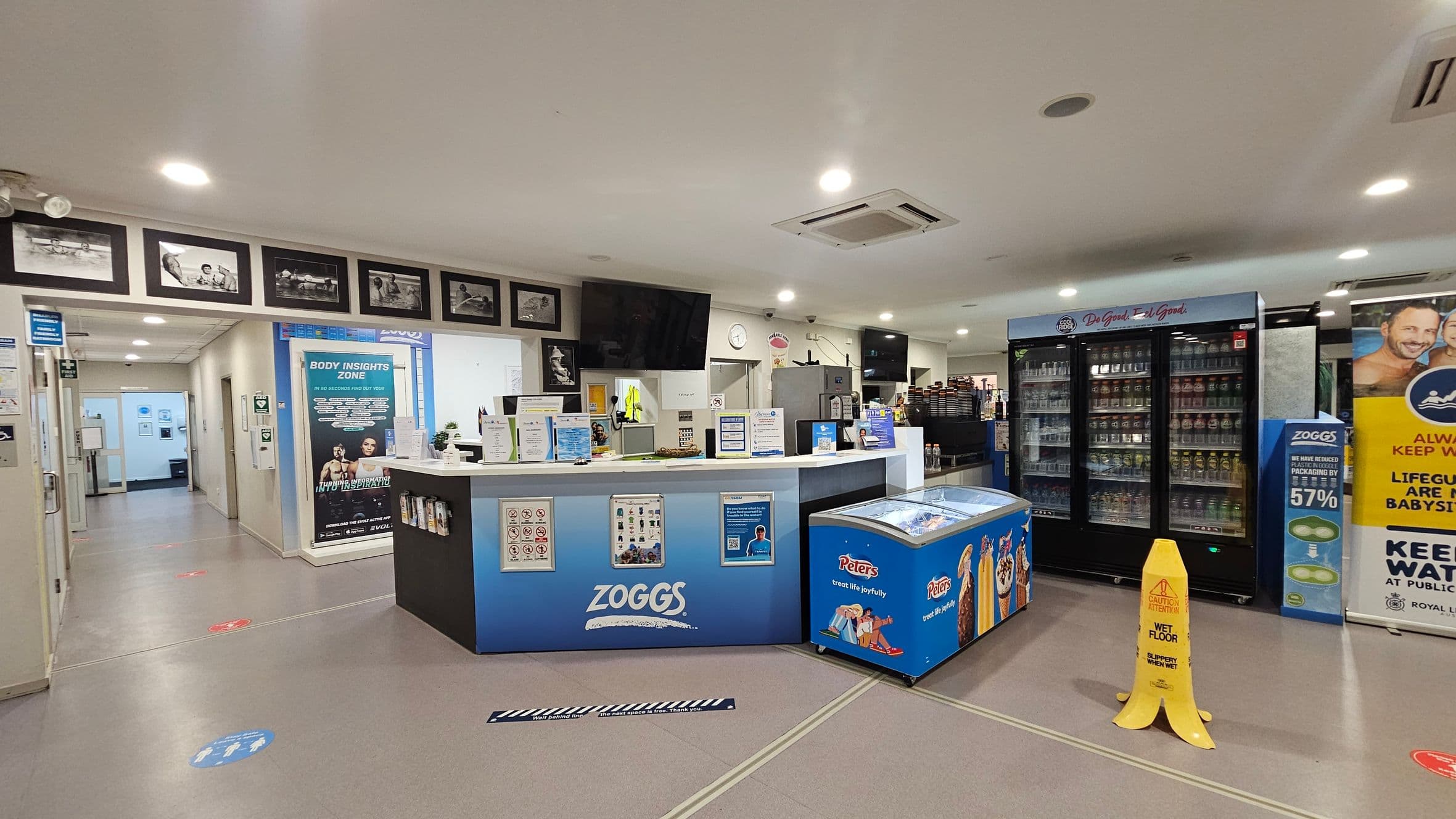 A reception area with a counter in the centre displaying various notices and a "Zoggs" logo. Behind the counter are two black screens and a clock on the wall. To the right, there is a large refrigerated display filled with drinks and a blue ice cream freezer with "Peters" branding. A yellow wet floor sign is on the floor. On the left, a hallway leads to other rooms, with framed photos on the wall above. The ceiling is white with recessed lighting.