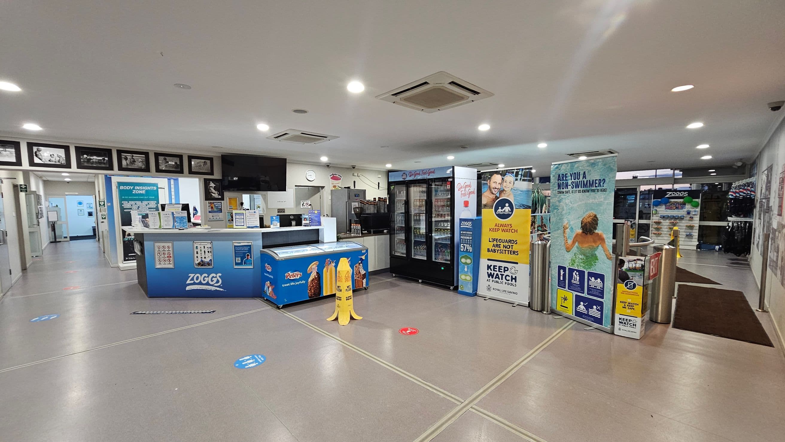 A reception area with a counter on the left displaying signs and a Zoggs-branded freezer. Behind the counter are framed photos and a wall-mounted TV. In the centre, a wet floor sign is visible. On the right, there are vending machines and promotional banners about pool safety. The floor is grey with social distancing markers, and the ceiling is white with recessed lighting. In the background, there is a hallway leading to other areas.