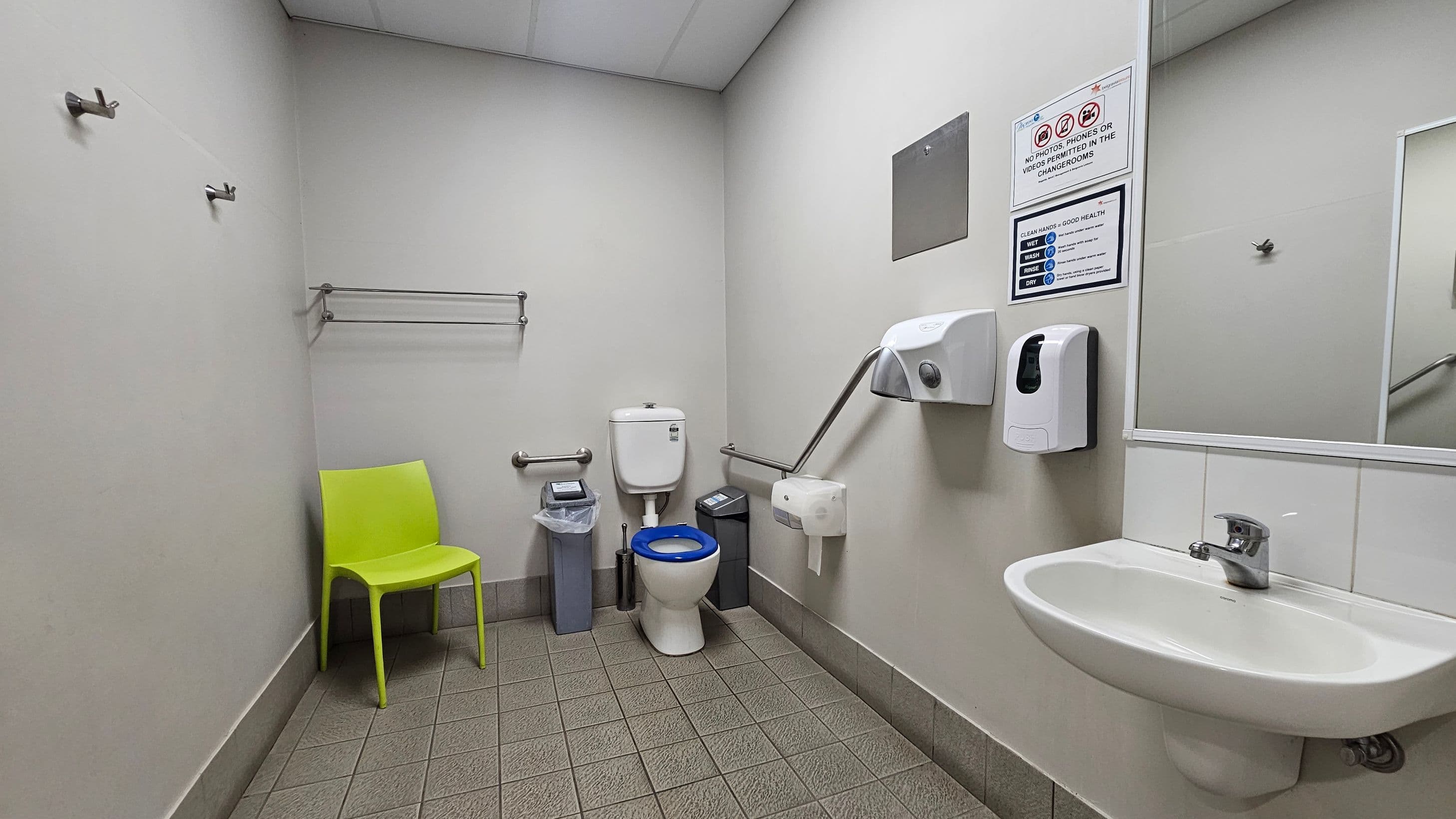 A public restroom with a green chair on the left, a toilet with a blue seat in the centre, and a small bin nearby. The walls have a towel rack and hooks. On the right, there's a sink with a soap dispenser and a hand dryer above it. A mirror is mounted on the wall, and there are informational signs above the sink. The floor is tiled in a light green pattern, and the ceiling is plain with recessed lighting.