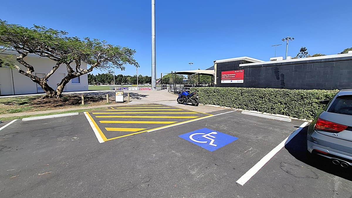 On the left, a large tree provides shade over a grassy area, with a few wooden posts visible nearby. A parking space is marked with yellow lines, including a blue symbol for disabled parking. A blue motorcycle is parked in this space. To the right, a building with a black exterior features a sign indicating "Athletics Queensland." The ground is asphalt, clearly marked, and the area appears tidy, with a low hedge bordering the parking lot.