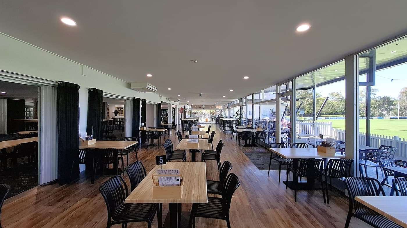 On the left, a series of black curtains frame an entrance, leading to a more private area. Several wooden tables with black chairs are arranged in the foreground, with a small menu holder on one table. In the centre, the floor is made of polished wooden boards, creating a warm appearance. To the right, large glass windows allow light to flood the space, revealing outdoor seating and greenery beyond. The ceiling is white with recessed lighting.