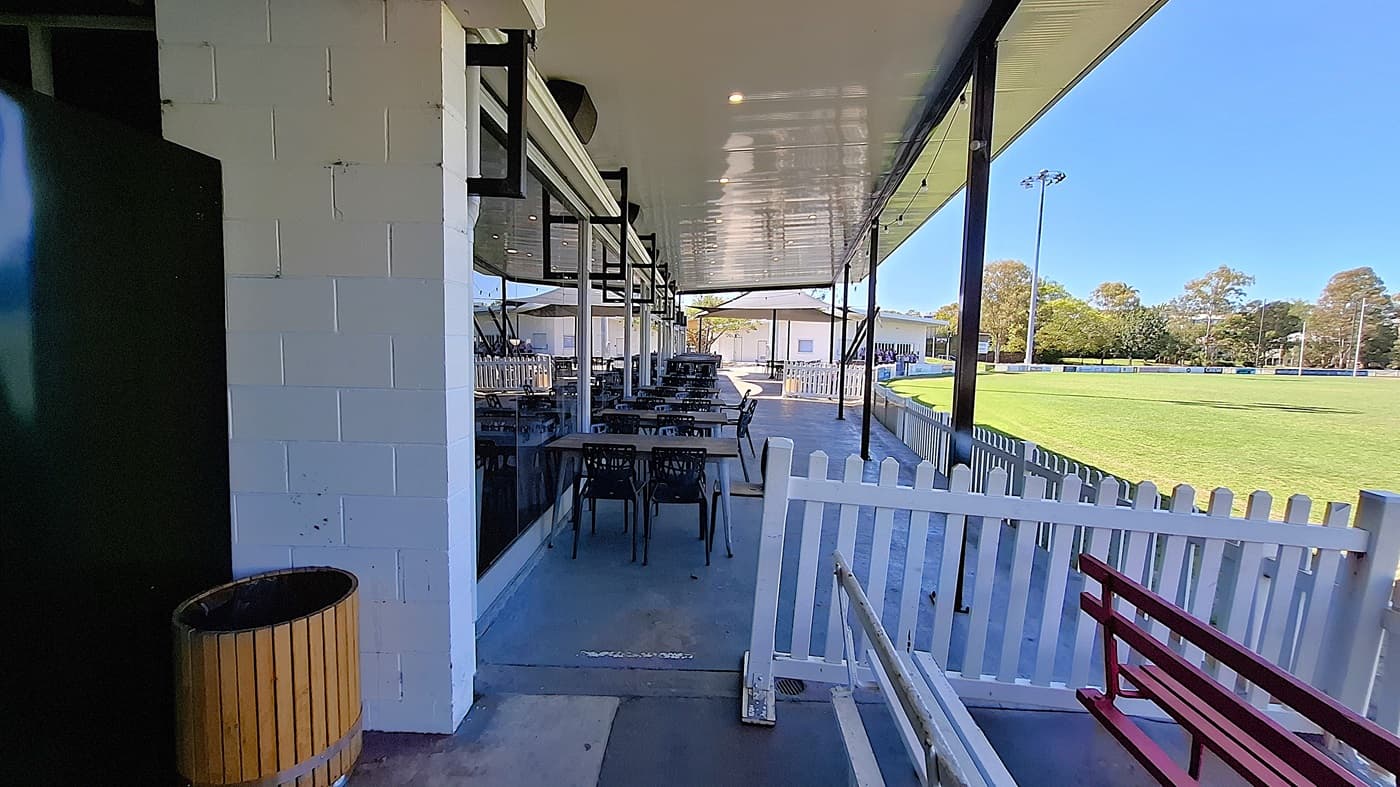 On the left, a green wall is visible next to a wooden bin, while a series of tables and chairs are arranged along the covered area. The floor is smooth and grey, extending towards the right. In the background, a white picket fence borders a green field, with several buildings visible beyond it. To the right, a red bench is positioned against the fence. The ceiling is flat and white, with overhead lights.