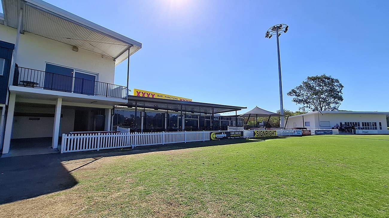On the left, a two-storey building features a balcony with black railings and large windows. Below, a covered area with glass panels provides seating. The floor is a mix of grass and concrete, leading to a well-maintained green field in the foreground. To the right, another building with white walls and large windows is visible, along with a tall light pole. The ceiling of the covered area is flat and extends outwards.  A white picket fence runs along the front.