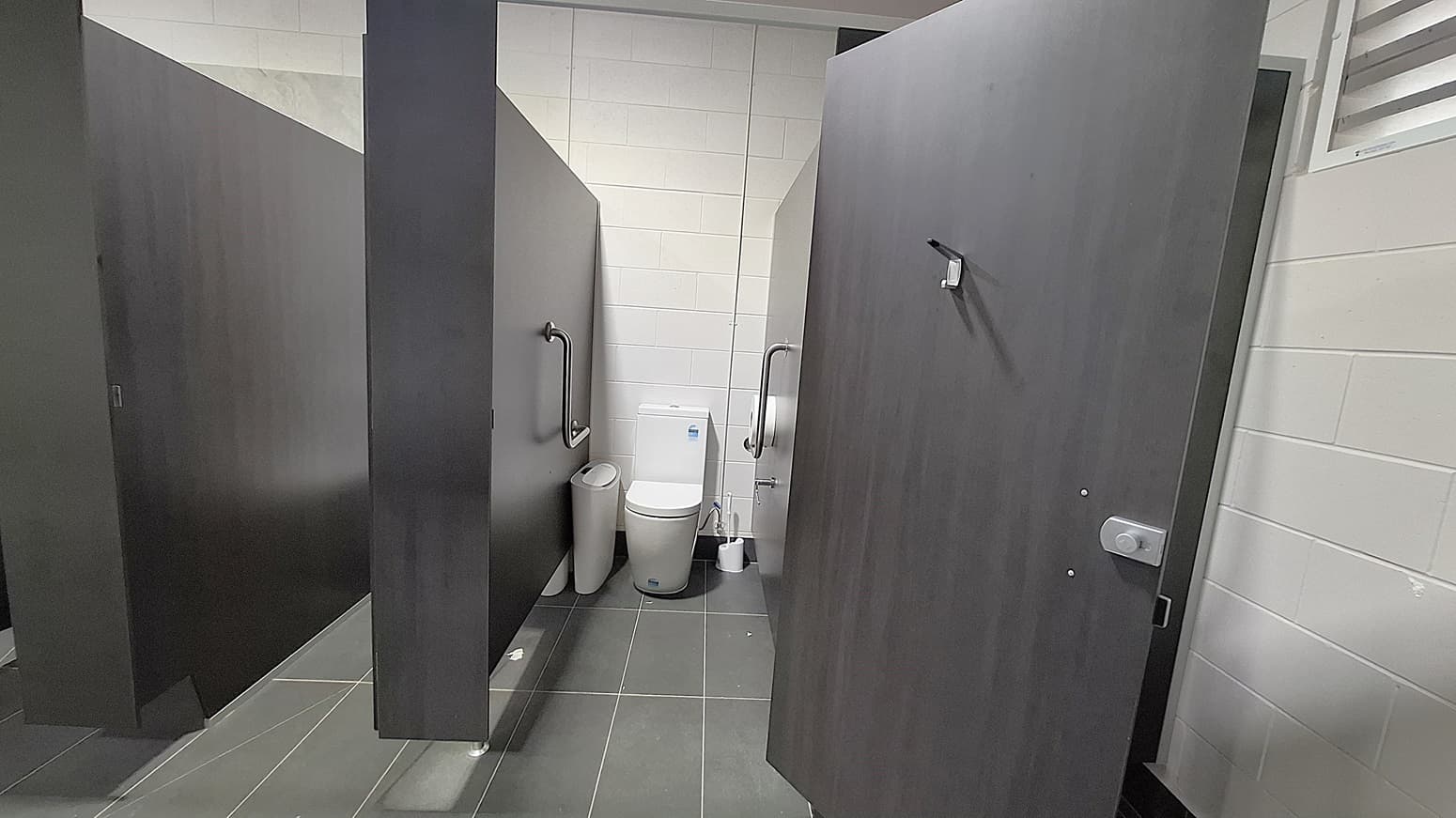 On the left, a dark wooden cubicle door stands closed, with a grab rail mounted beside it. Adjacent to it, a white toilet is positioned against the wall, with a small bin nearby. The floor is dark grey tiles, extending throughout the space. To the right, another cubicle door is slightly ajar, revealing a similar setup. The ceiling is flat and white, with ventilation grilles visible, providing a clean and functional environment.