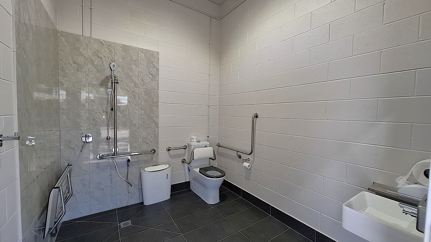 On the left, a shower area features a wall of grey tiles with a handheld showerhead mounted above. A grab rail is installed beside the shower. Adjacent to it, a white toilet is positioned with a roll of toilet paper mounted on the wall. The floor is dark grey tiles, extending throughout the space. To the right, a small sink is mounted on the wall, with a paper towel dispenser nearby. The ceiling is flat and white, with exposed pipes running along it.