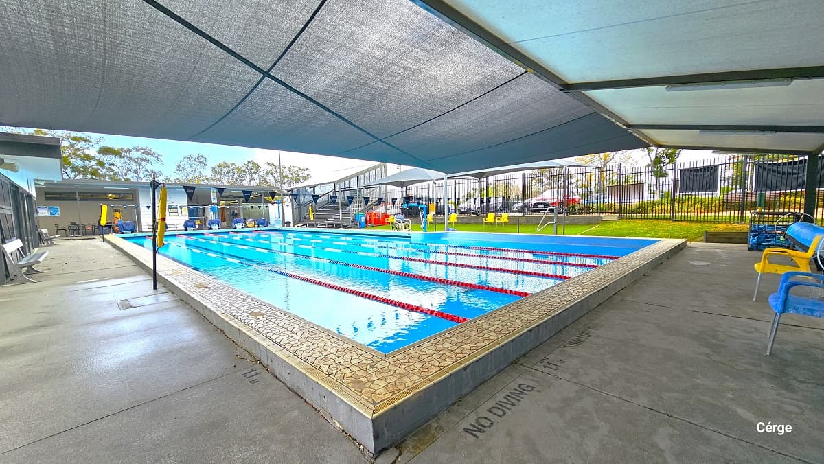 25m with 6 lanes outdoor aquatic pool at Mudgeeraba Aquatic Centre. The pool lanes are separated by red, white, and blue floating rope lanes. Two strings of triangular blue flags are tied around the opposite poles with a yellow flotation device. Seen at the back of this image are diving platforms covered with a blue cloth at the start of each pool lane, and to the right is multi-tiered seating. Yellow, blue, black, and grey plastic chairs are available all throughout the area.