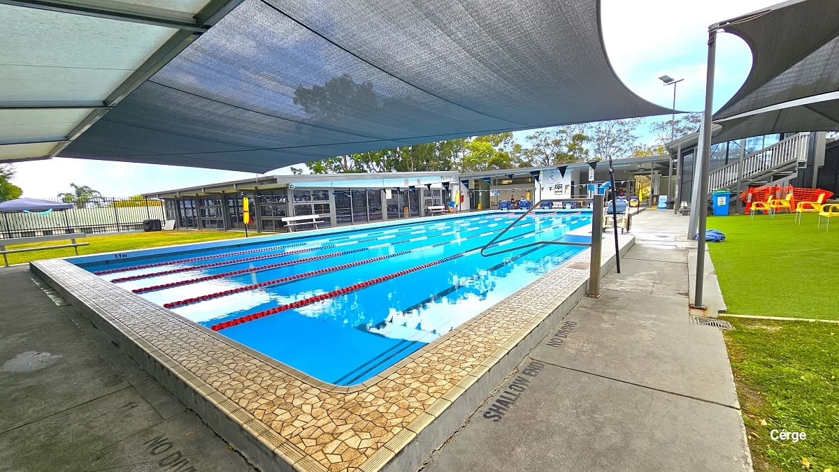 Shallow end of the 25m at Mudgeeraba Aquatic Centre, near the two black sunshades with yellow plastic chairs. It has no diving mark on the poolside. Opposite this side is a large open space with Bermuda grass, a blue tent, and a black fence. Seen above the entire facility are large trees with abundant foliage.