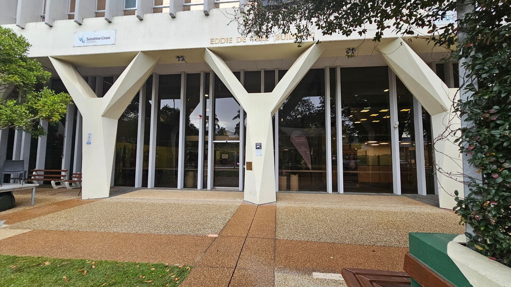 The image shows the entrance of the Eddie De Vere Building at Sunshine Coast Council. The facade features large glass windows and distinctive Y-shaped columns. A sign with the council's logo is visible above the entrance. The ground is covered with a textured, multicoloured surface leading to the doors. On the left, there is a bench and a tree, while on the right, a wall with climbing plants is visible. Inside, ceiling lights and some interior furnishings can be seen through the glass.