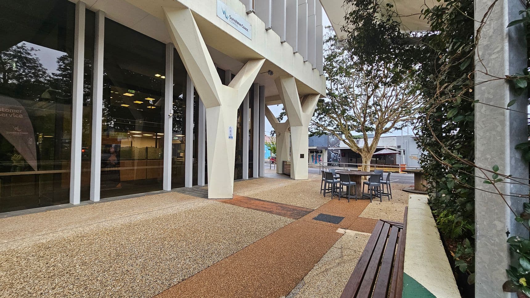 A modern building with large glass windows and angular white supports on the left. The ground is covered with a textured, speckled surface in shades of beige and brown. In the centre, there is a small outdoor seating area with tables and chairs. On the right, a wall is partially covered with green climbing plants. A tree is visible in the background, along with other buildings. A wooden bench is in the foreground on the right.