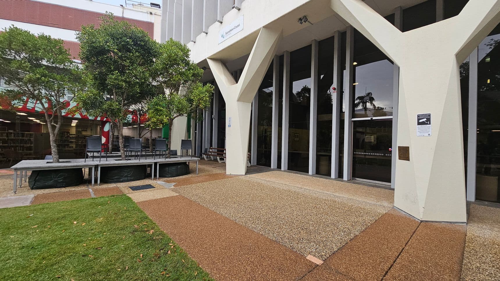A building entrance with large glass doors and distinctive Y-shaped columns. To the left, there are several trees planted in raised platforms with chairs placed around them. The ground is a mix of grass and textured paving. In the background, a building with horizontal red and white stripes is visible. A sign is mounted above the entrance, and a security notice is posted on one of the columns.