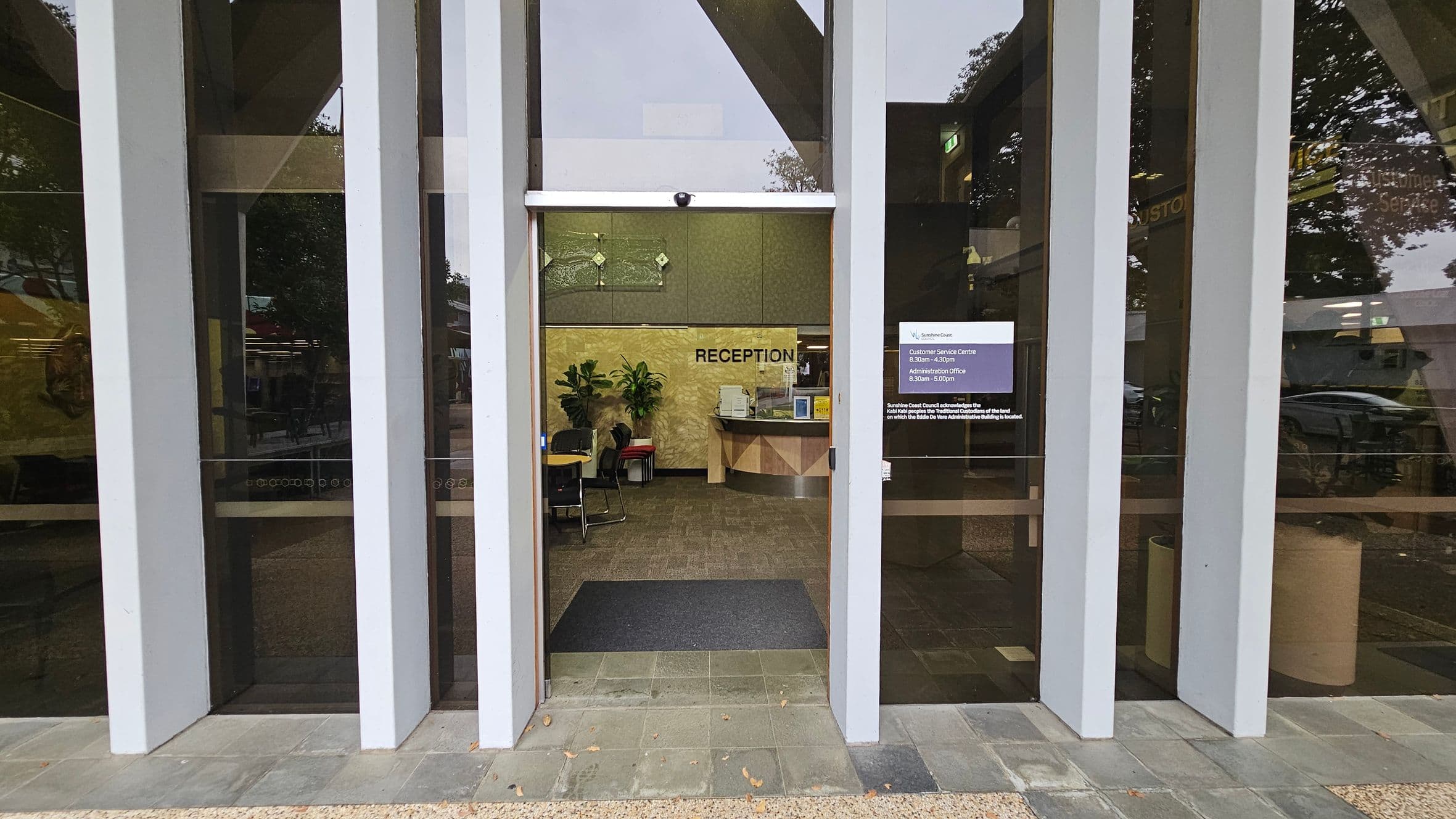 A glass entrance with vertical white beams leads to a reception area. Inside, a counter with a computer and office supplies is visible. The word "RECEPTION" is displayed on the back wall. Potted plants and chairs are arranged near the counter. A sign on the right glass panel provides customer service information. The floor outside is tiled, transitioning to carpet inside. Trees and reflections are visible through the glass.