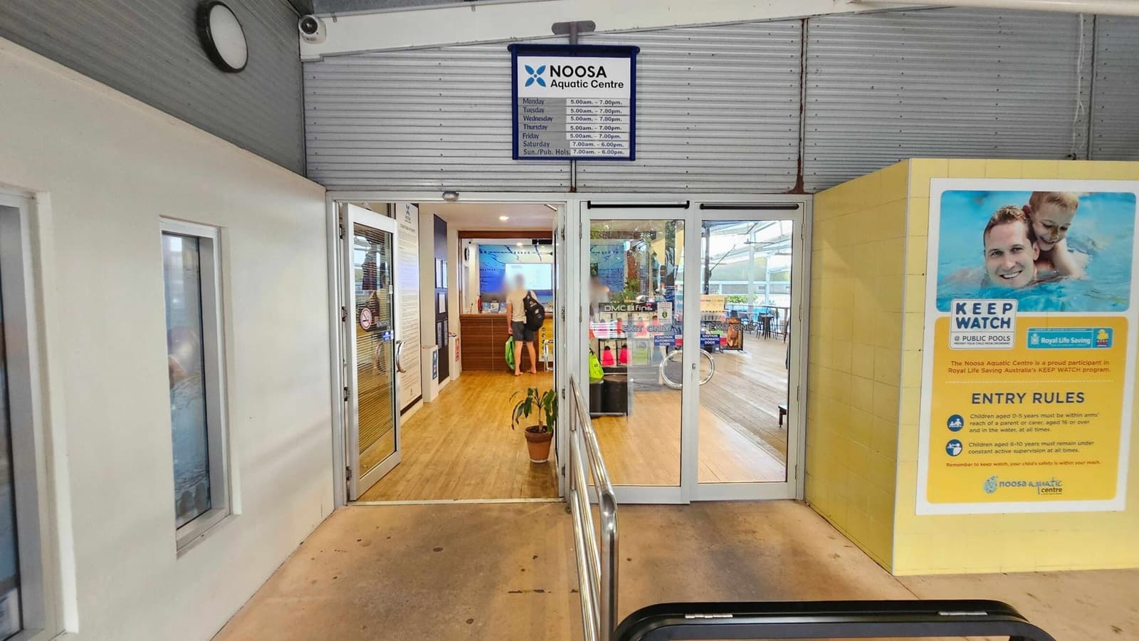 Entrance to the Noosa Aquatic Centre with glass doors in the centre. Above the doors, a sign displays the centre's name and opening hours. To the right, a large poster outlines pool safety rules with an image of a man and child swimming. Inside, a person stands near a wooden reception desk, and various swimming gear is visible. The floor is wooden, and a potted plant is placed near the entrance. The ceiling is light-coloured with a clock and security camera.