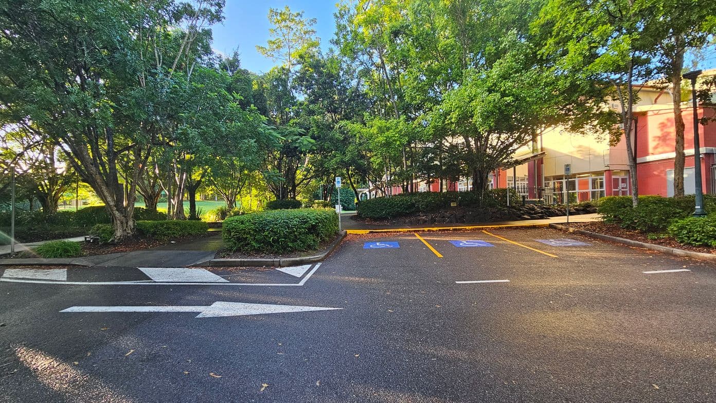 A parking area with marked disabled spaces is bordered by lush green trees and shrubs. The ground is asphalt with clear white directional arrows and blue disabled parking symbols. To the right, a red brick building with large windows is partially visible. A pathway leads through the greenery, and a lamppost stands near the building. The area feels open with a mix of natural and built elements.