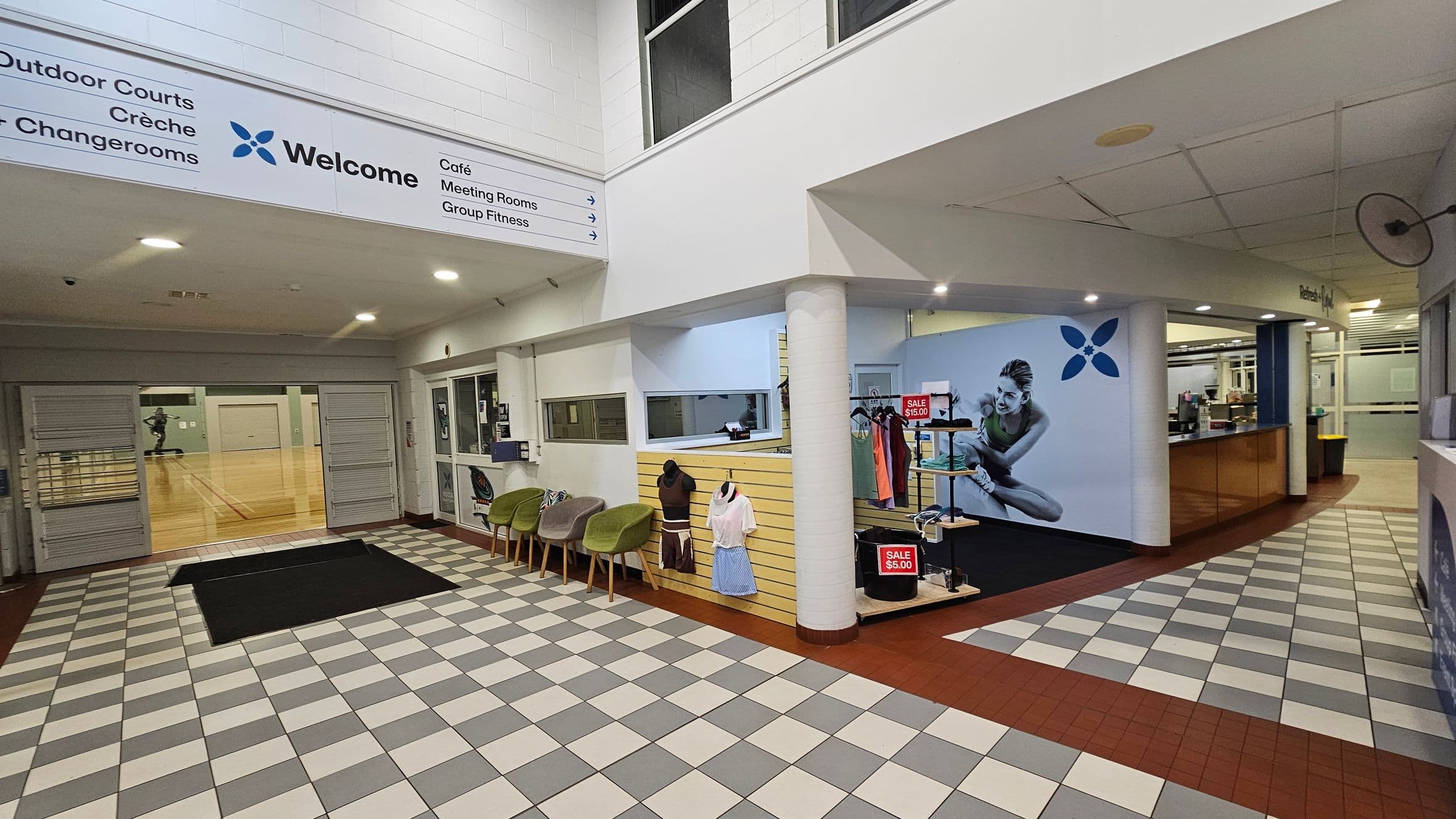 The image shows an indoor sports facility entrance with a tiled floor in a checkerboard pattern of grey and white. To the left, a large sign directs to various areas like outdoor courts and a café. A reception area with a counter and a display of clothing on sale is visible on the right. The space is enclosed with white walls and a low ceiling, featuring a prominent column. A ramp leads into a gymnasium with wooden flooring.