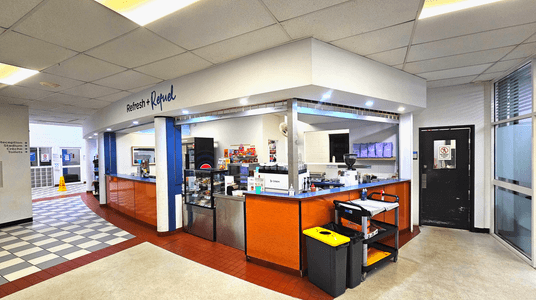 The image shows a refreshment kiosk with a sign reading 'Refresh + Refuel' above it. The kiosk features a blue counter with orange panels and a variety of snacks and drinks displayed. To the left, a tiled floor transitions from red to a black and white checkered pattern leading to a corridor with signage for reception, stadium, crèche, and toilets. A black door with a window is on the right, next to a glass panel.