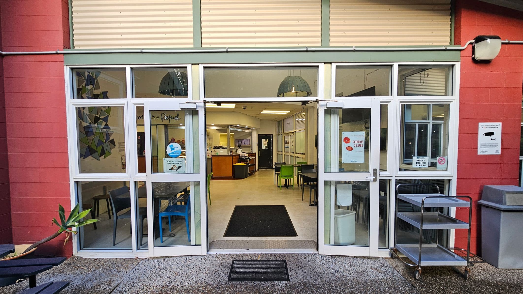 The image shows a glass double-door entrance to a building with a red brick exterior. Inside, there are tables and chairs in various colours, including green and blue. A black mat lies at the entrance, and a metal trolley with three shelves is positioned to the right. The floor transitions from outdoor concrete to indoor tiles. Posters and signs are visible on the doors and walls, indicating a functional space, possibly a café or dining area.