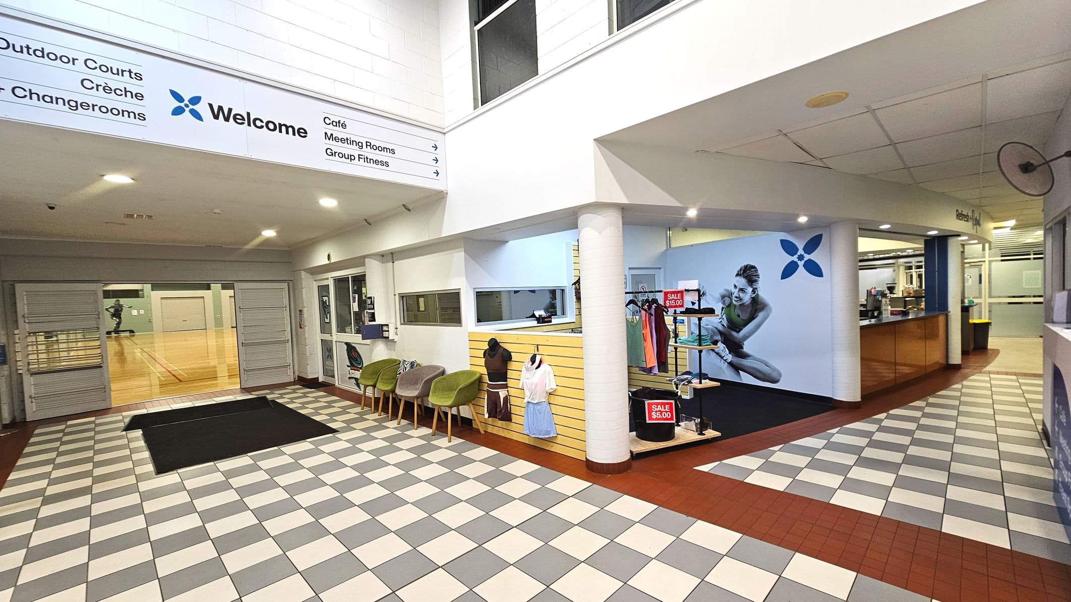 The image shows an indoor area with a tiled floor featuring alternating grey and white squares. On the left, there is a large entrance with glass doors leading to a wooden-floored sports court. Above, a sign lists facilities like outdoor courts and a café. To the right, a reception area with a counter and a wall display of sportswear is visible. The space is enclosed with white walls and a ceiling with recessed lighting.