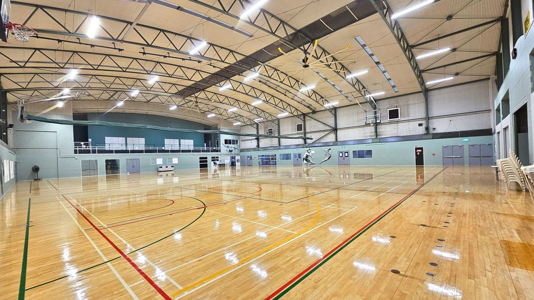 A spacious indoor sports hall with a polished wooden floor marked with various coloured lines for different sports. The ceiling is high with exposed metal trusses and bright overhead lighting. Basketball hoops are mounted on the walls, and there are several doors and windows along the perimeter. A stack of chairs is visible on the right side. The walls are painted in light blue and grey tones, giving a modern appearance.