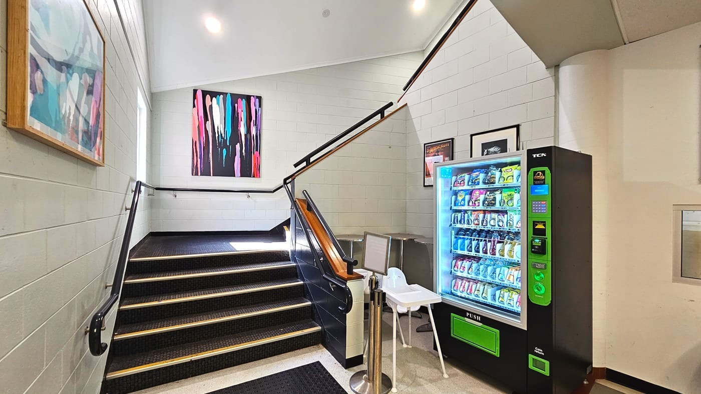 A stairwell with black steps and yellow safety edges leads upwards, flanked by white walls adorned with colourful abstract paintings. A black handrail runs along the stairs. To the right, a brightly lit vending machine filled with snacks stands next to a white high chair. The floor transitions from a black mat to a light-coloured tile. The space feels enclosed with a low ceiling and walls on all sides.