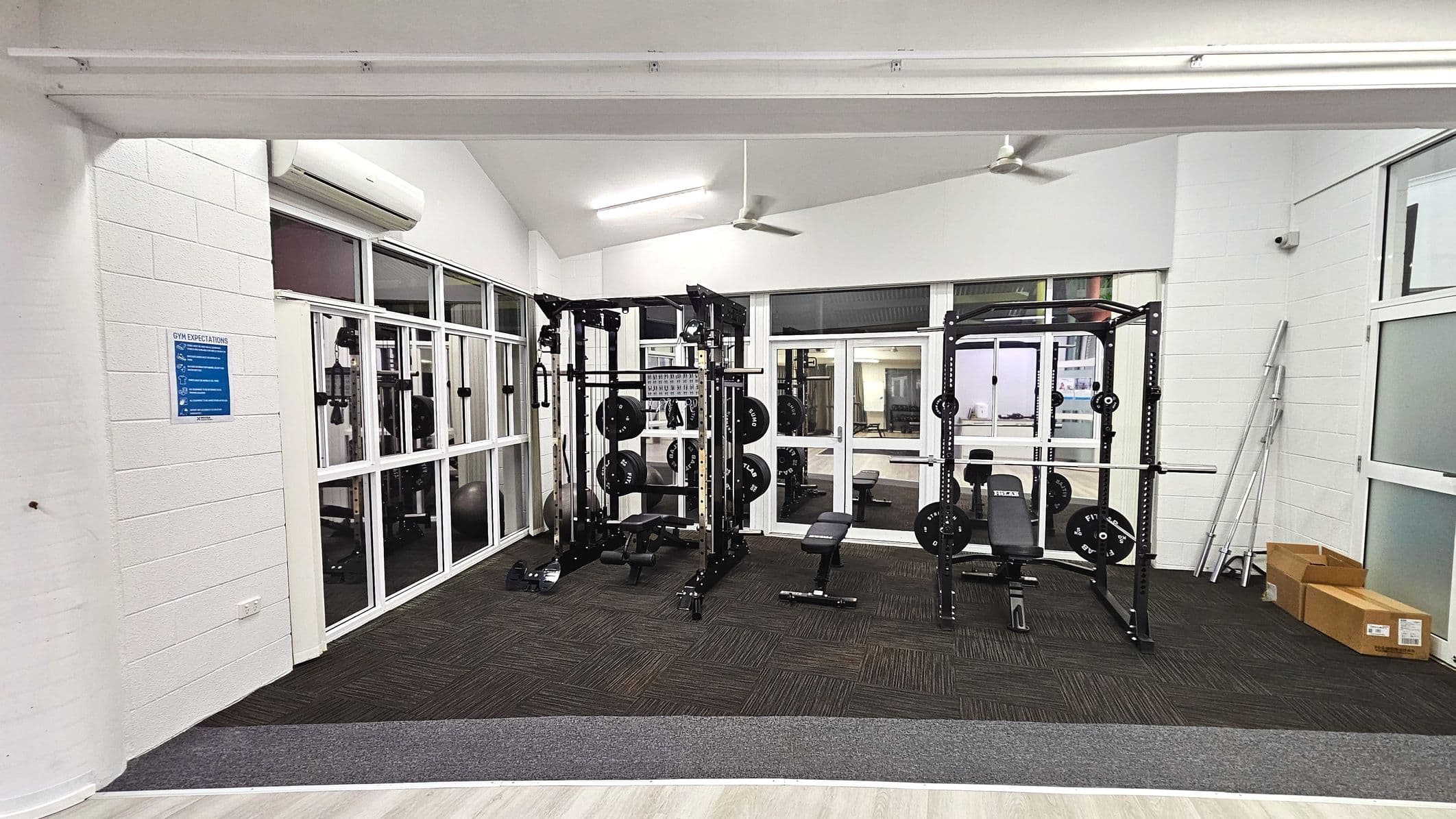 A gym area with two weightlifting racks equipped with barbells and weights, positioned on a dark carpeted floor. The space is enclosed by white walls and large glass windows, providing a view into another room. A blue sign is mounted on the left wall. Ceiling fans and fluorescent lights are visible above. Cardboard boxes and metal poles are stacked in the right corner. The floor transitions from carpet to a lighter material at the entrance.