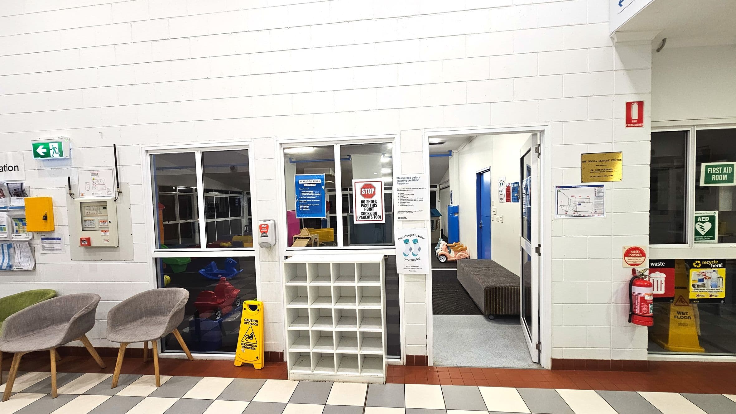 The image shows an indoor area with white brick walls and a tiled floor in shades of green, white, and brown. On the left, there are two grey chairs and a wall-mounted brochure holder. A green exit sign and a yellow caution wet floor sign are visible. In the centre, a shoe cubby and a window with signs about shoe removal are present. To the right, a doorway leads to a room with a blue door and children's toys. A fire extinguisher and first aid signage are also visible.
