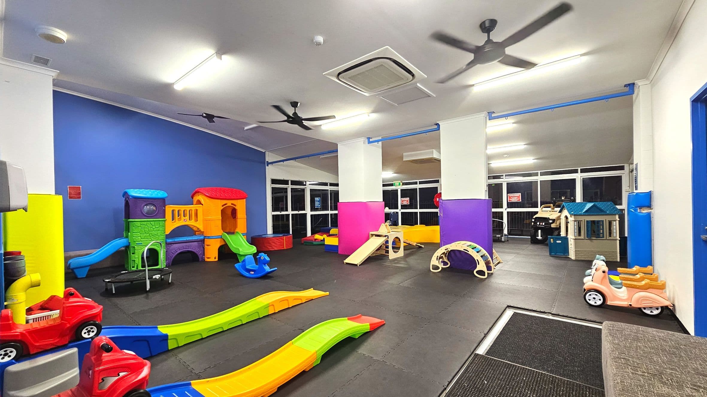 A brightly coloured indoor play area with various plastic play structures. On the left, a large multi-coloured playhouse with slides and tunnels is set against a blue wall. The floor is covered with interlocking black mats. In the centre, small slides and a rocking horse are visible. To the right, a playhouse and a small arch tunnel are near large windows. Ceiling fans and fluorescent lights are overhead.