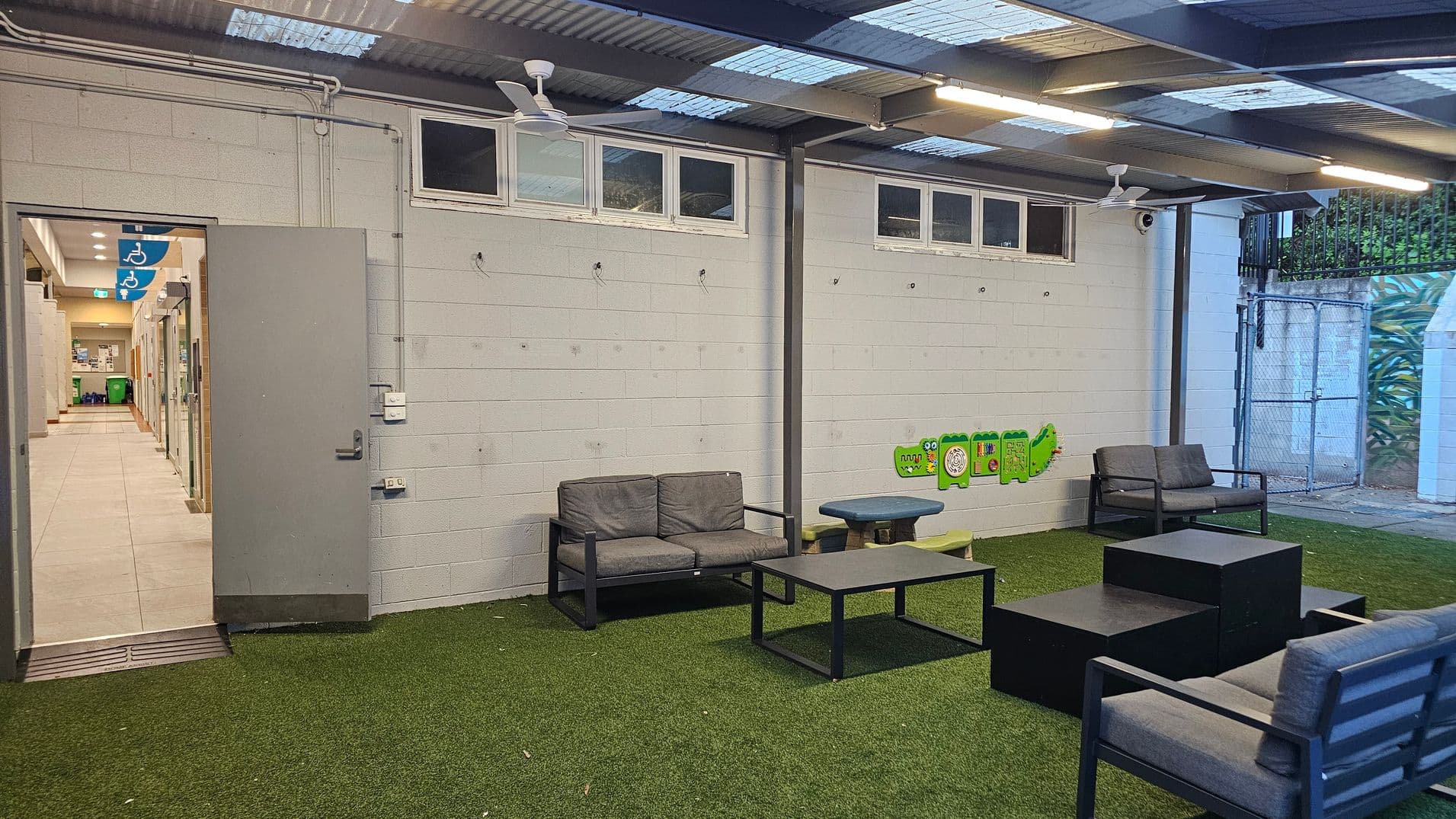 An indoor area with artificial grass flooring features several grey cushioned chairs and black tables arranged in a seating area. The left side has an open door leading to a corridor with tiled flooring and visible signage for accessible facilities. The walls are light-coloured brick with small high windows and a ceiling with exposed beams and fans. A chain-link gate is visible in the background.