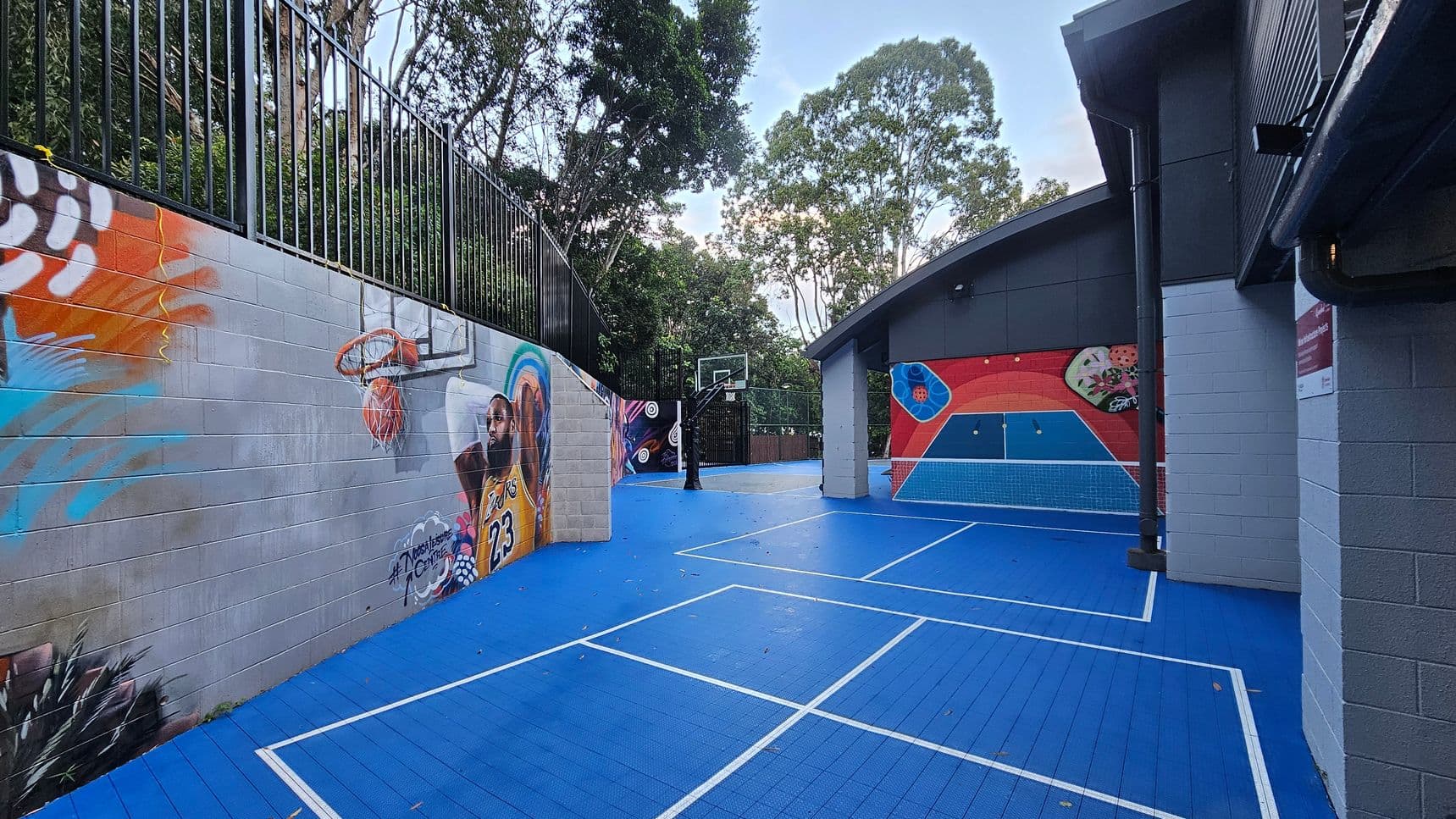 A vibrant outdoor sports court with a bright blue surface and white lines, featuring a basketball hoop at one end. The left side is bordered by a mural-covered wall with colourful graffiti and a metal fence above. To the right, a building with grey walls and a sloped roof displays more murals. Tall trees surround the area, providing a natural backdrop. The space feels enclosed by the wall and building.