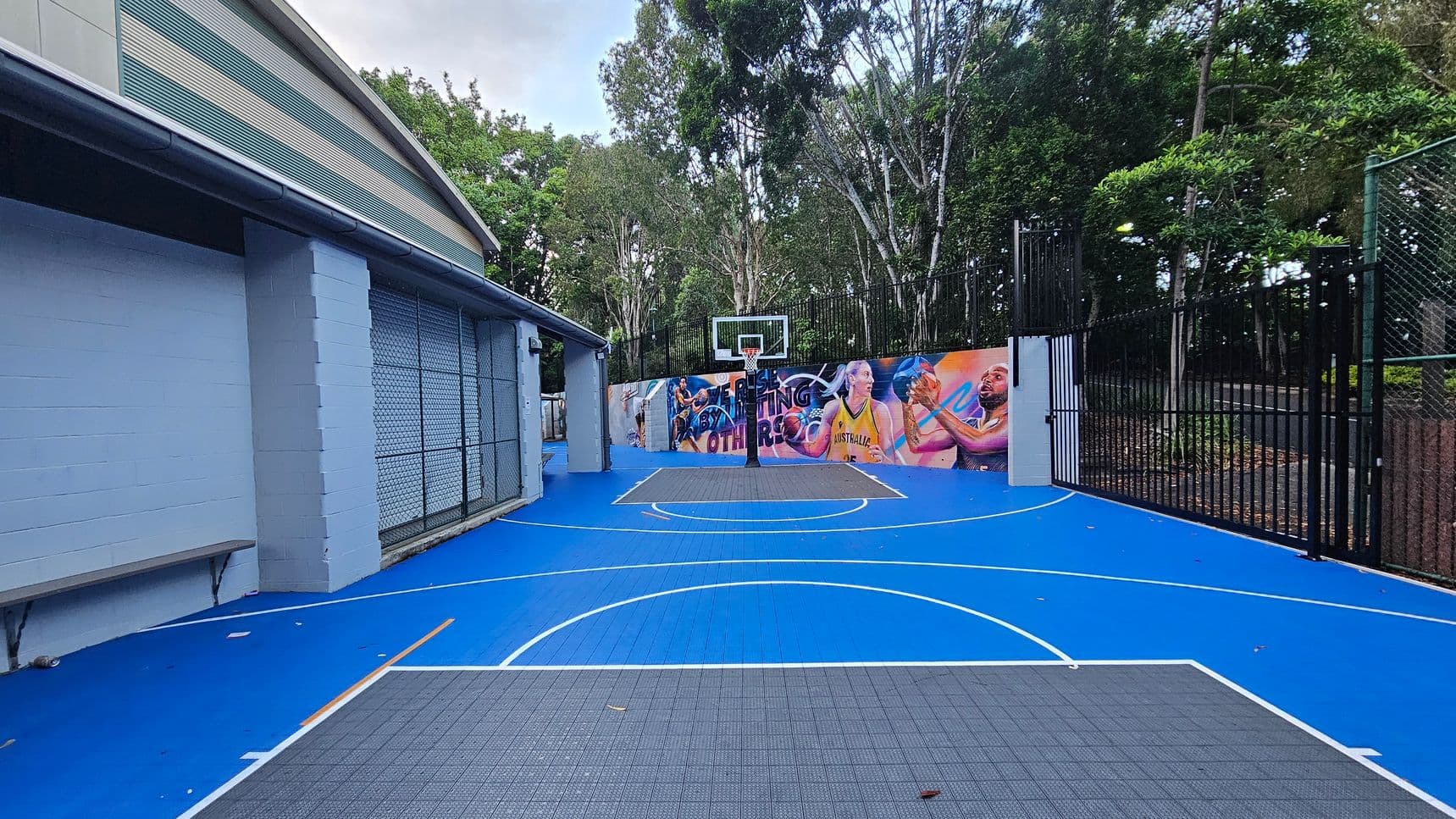 An outdoor basketball court with a vibrant blue surface and white lines, featuring a central hoop with a backboard. The court is bordered by a colourful mural depicting athletes, and a black metal fence encloses the area. To the left, a building with light blue walls and a metal bench is visible. The court is surrounded by tall trees, providing a natural backdrop.