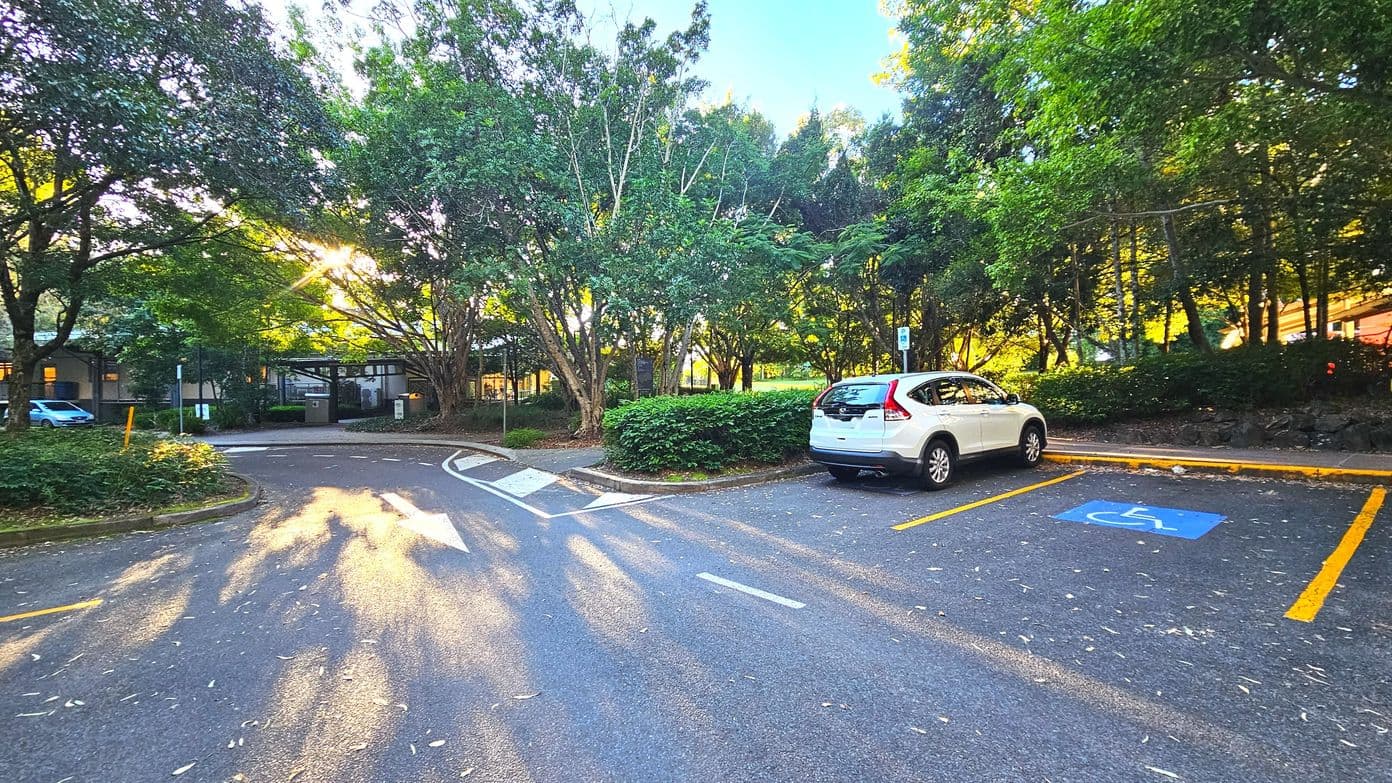A car park area with a white vehicle parked in a designated disabled parking space marked by blue paint and a wheelchair symbol. The area is surrounded by lush green trees and bushes, creating a natural boundary. The asphalt surface is marked with white directional arrows and yellow lines. In the background, a building is partially visible through the trees, and sunlight filters through the foliage, casting shadows on the ground.