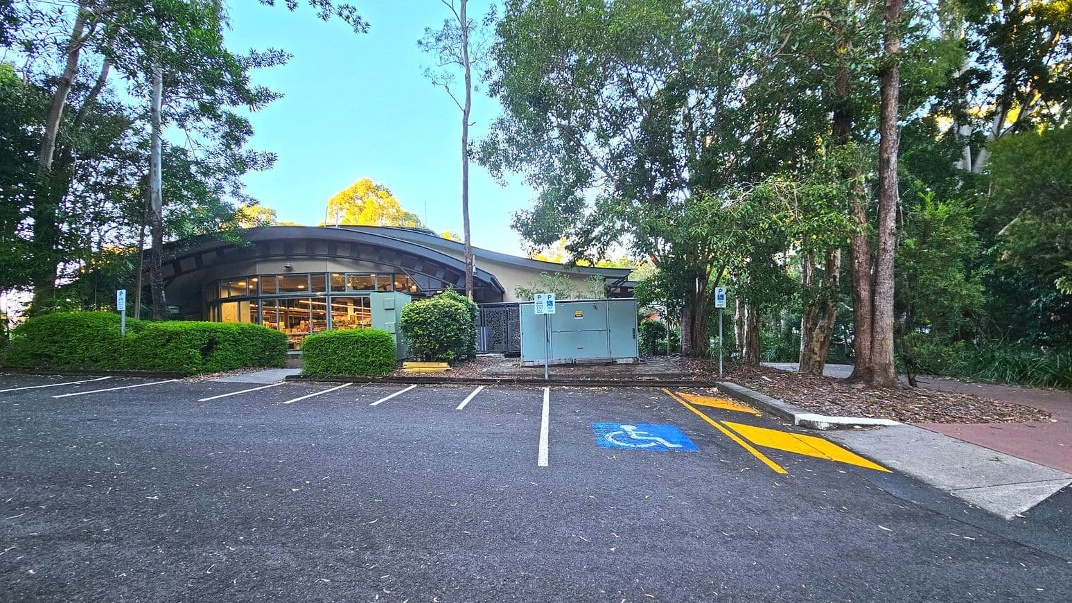 A curved building with large glass windows is partially obscured by trees and bushes. In front, there is a parking area with marked spaces, including a blue disabled parking spot with a yellow access zone. The ground is asphalt, transitioning to a concrete path on the right. The area is surrounded by tall trees, creating a semi-enclosed feel. Signs indicate parking regulations.