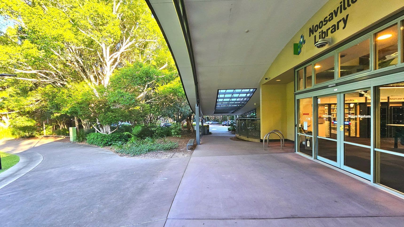 A library entrance with large glass doors and a sign reading 'Noosaville Library' above. The building has a curved roof with a glass section allowing light through. To the left, a paved path curves alongside lush greenery and tall trees. A bike rack is near the entrance, and a bench is positioned along the path. The area feels open with a mix of concrete and natural surfaces.