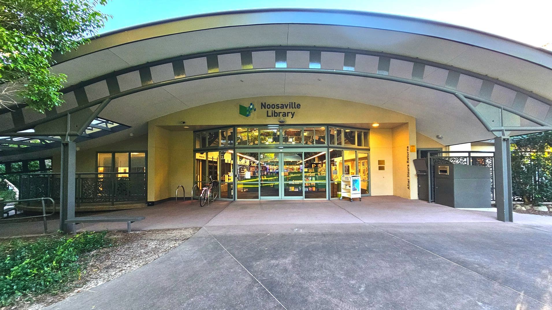 The entrance to Noosaville Library features a wide, curved metal awning with a geometric pattern, providing shade over the glass double doors. The library's name is displayed above the entrance. To the right, there's an after-hours book return slot. A bicycle rack is visible on the left, and the ground is paved with concrete. The area is bordered by greenery, creating a semi-enclosed feel.