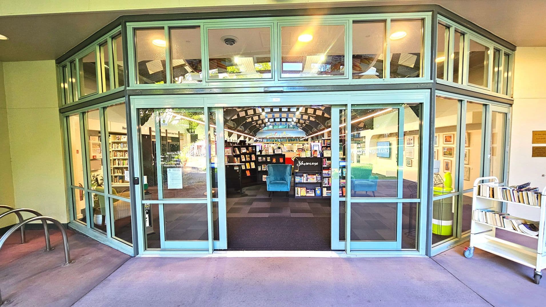 The image shows the entrance to a library with large glass doors framed by a series of rectangular windows above and on the sides. Inside, bookshelves are visible, along with a blue armchair and a sign reading 'Showcase'. The floor transitions from a purple exterior surface to a dark carpet inside. A book trolley is positioned to the right of the entrance. The space feels enclosed by the glass structure.