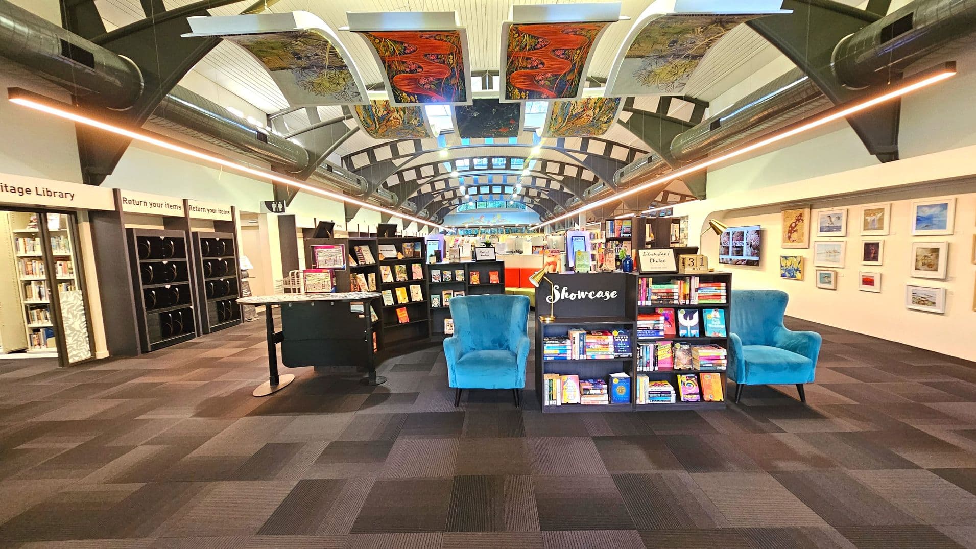 A modern library interior with a high arched ceiling featuring colourful artwork panels. The centre has a display area with a blue armchair flanked by bookshelves labelled 'Showcase'. To the left, there are book return slots and a sign for the Heritage Library. The floor is covered with dark patterned carpet tiles. The walls display framed artwork, and the space feels open with visible ductwork and lighting along the ceiling.