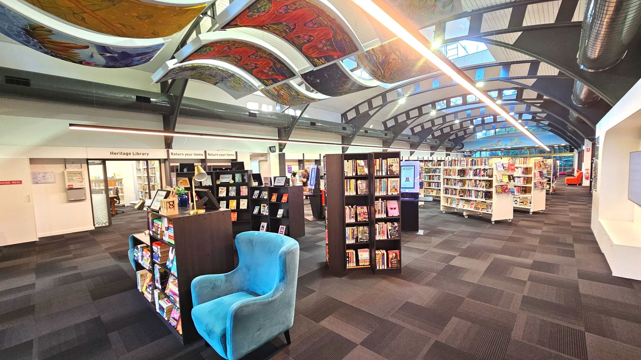 A spacious library interior with a high, arched ceiling featuring colourful, artistic panels. The floor is covered with dark, patterned carpet tiles. Bookshelves filled with books are arranged neatly, with a prominent blue armchair in the foreground. Signs for 'Heritage Library' and 'Return your items' are visible on the left. The space is open and well-lit, with large windows at the far end allowing natural light in.