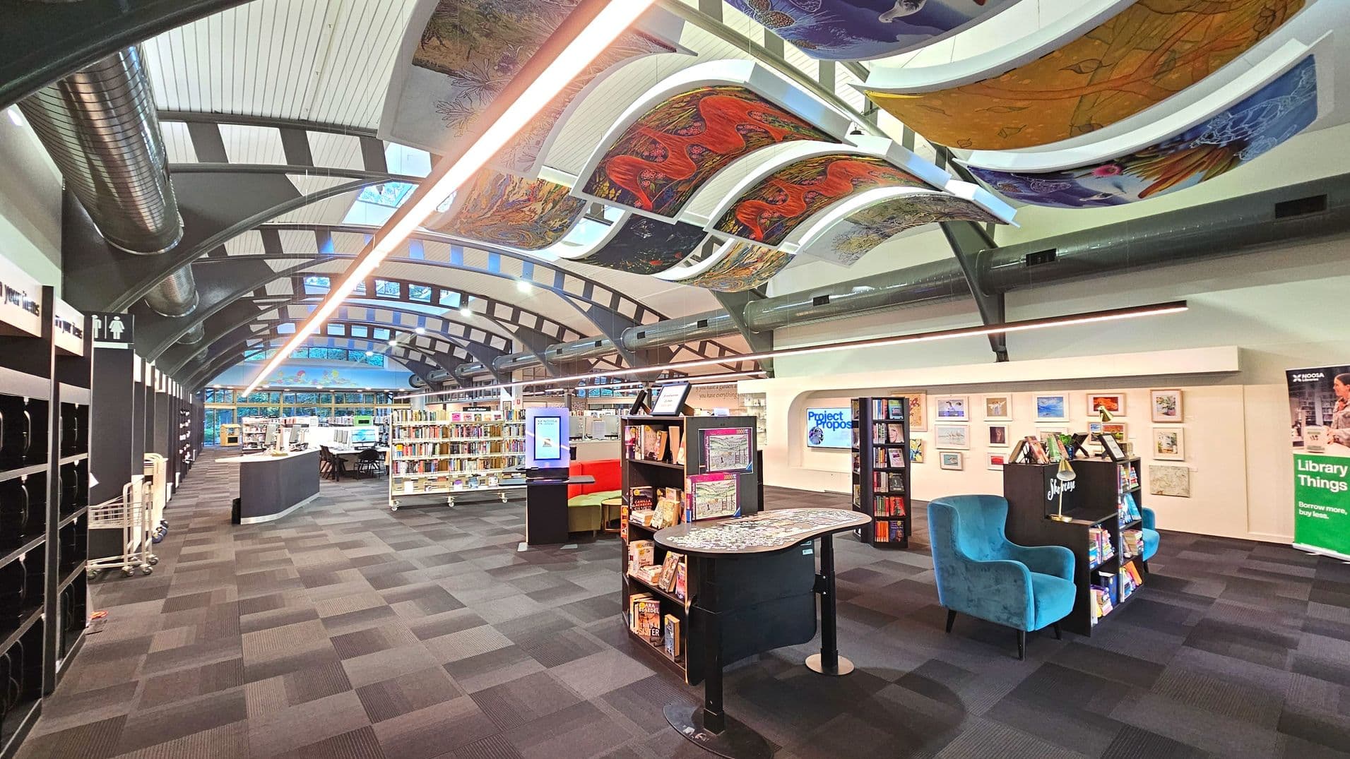 A modern library interior with a high, curved ceiling featuring colourful, artistic panels. The space is open with bookshelves lining the left and centre, and a seating area with blue armchairs on the right. A digital kiosk and a curved information desk are visible. The floor is covered with patterned carpet tiles. A sign for restrooms is on the left wall, and framed artwork decorates the right wall.