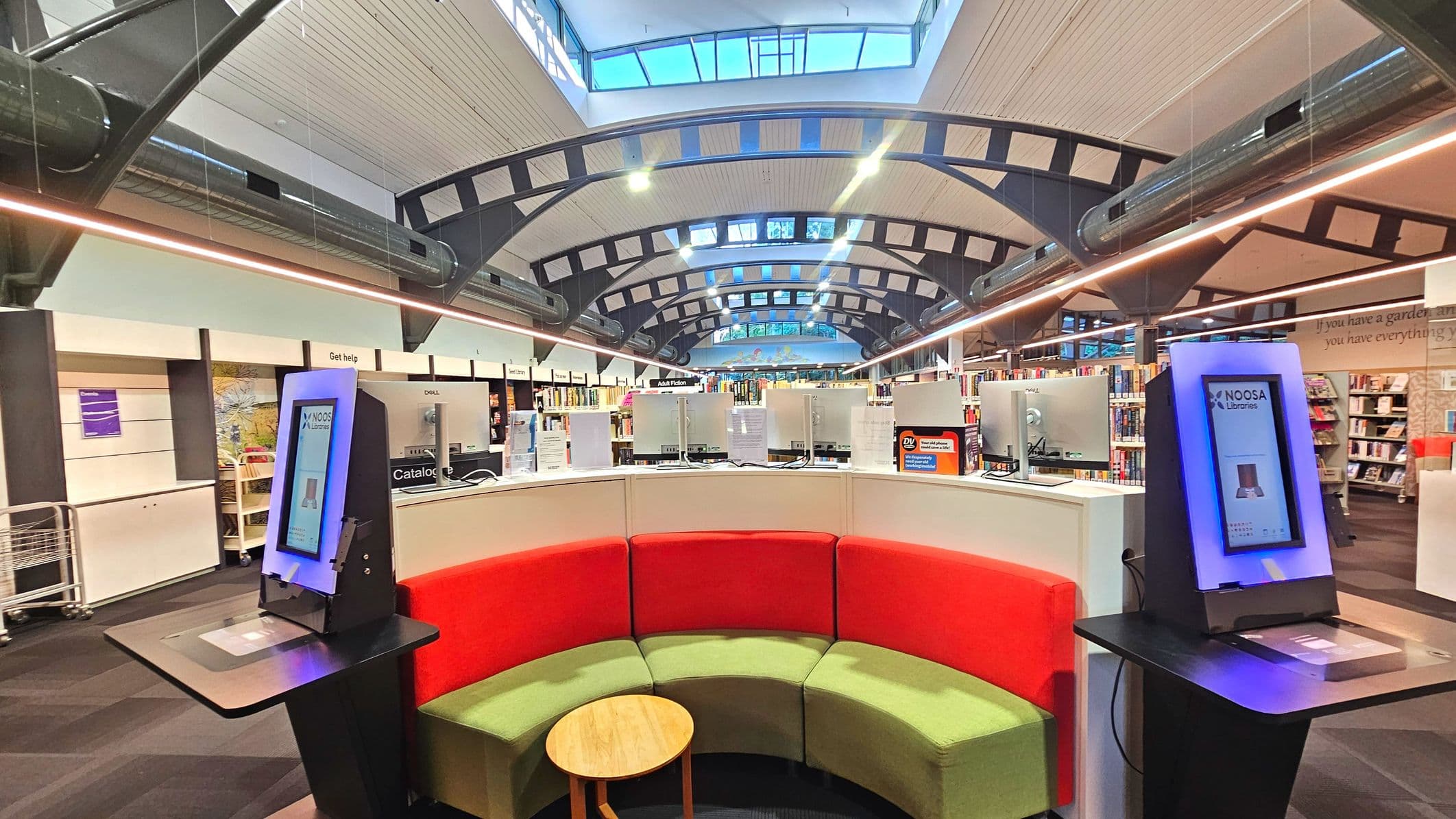 The interior of a modern library features a curved seating area with red and green cushions surrounding a small wooden table. Two digital kiosks with screens are positioned on either side of the seating. Above, a high ceiling with exposed beams and skylights allows natural light to illuminate the space. Bookshelves line the background, and a 'Get help' sign is visible on the left. The floor is carpeted in a dark colour.