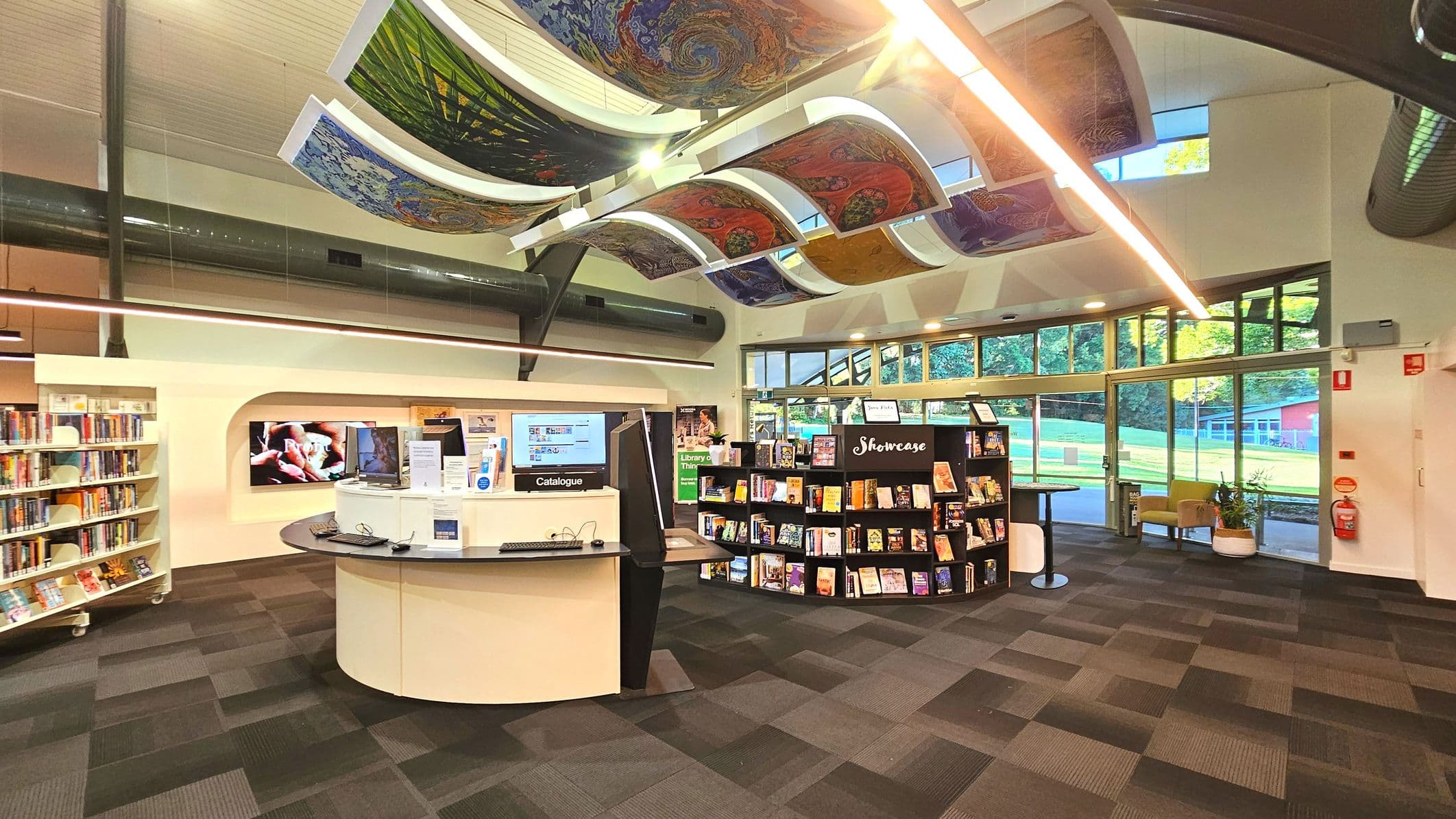 A modern library interior with a curved ceiling featuring colourful, artistic panels. The space includes a central circular information desk with computers labelled 'Catalogue'. To the left, bookshelves are filled with books, and to the right, a 'Showcase' display holds various items. Large windows line the back wall, offering views of greenery outside. The floor is covered with patterned carpet tiles, and a fire extinguisher is mounted on the right wall.