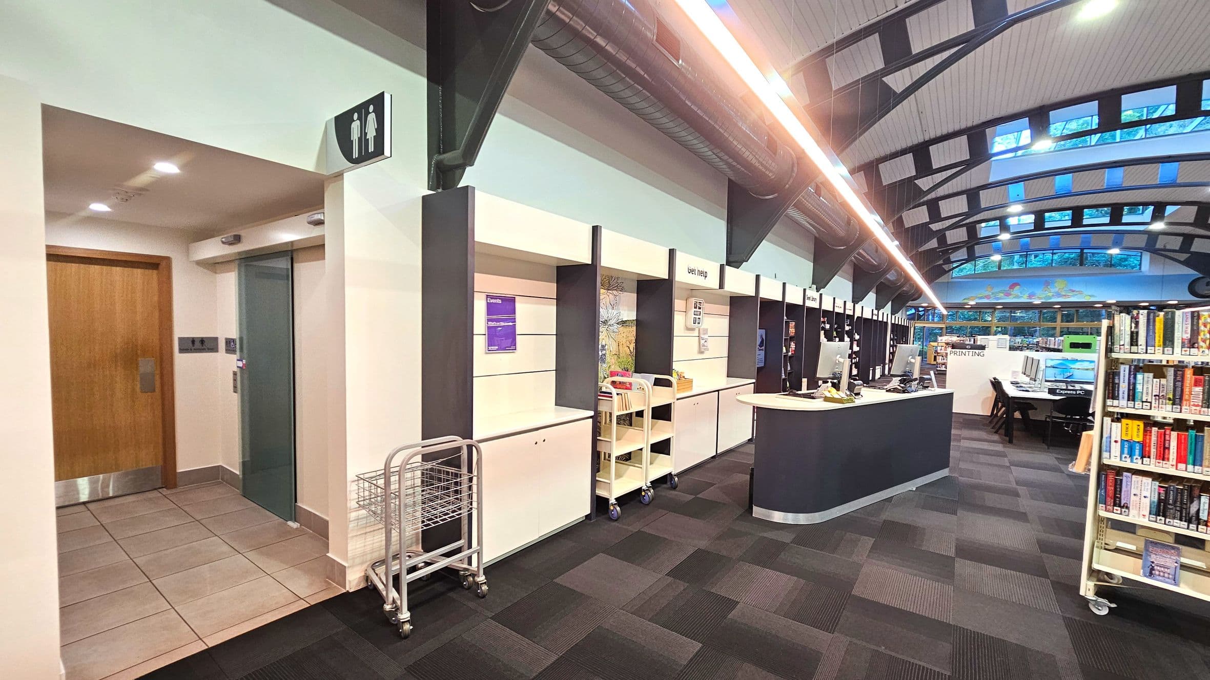 The image shows an interior space with a modern design. On the left, there's a wooden door next to a lift with a metallic door, under a ceiling with recessed lighting. A sign with male and female symbols indicates nearby restrooms. The floor transitions from light tiles to dark carpet tiles. To the right, there are service counters with signage like 'Print Help', and a row of computers. Bookshelves are visible in the background.