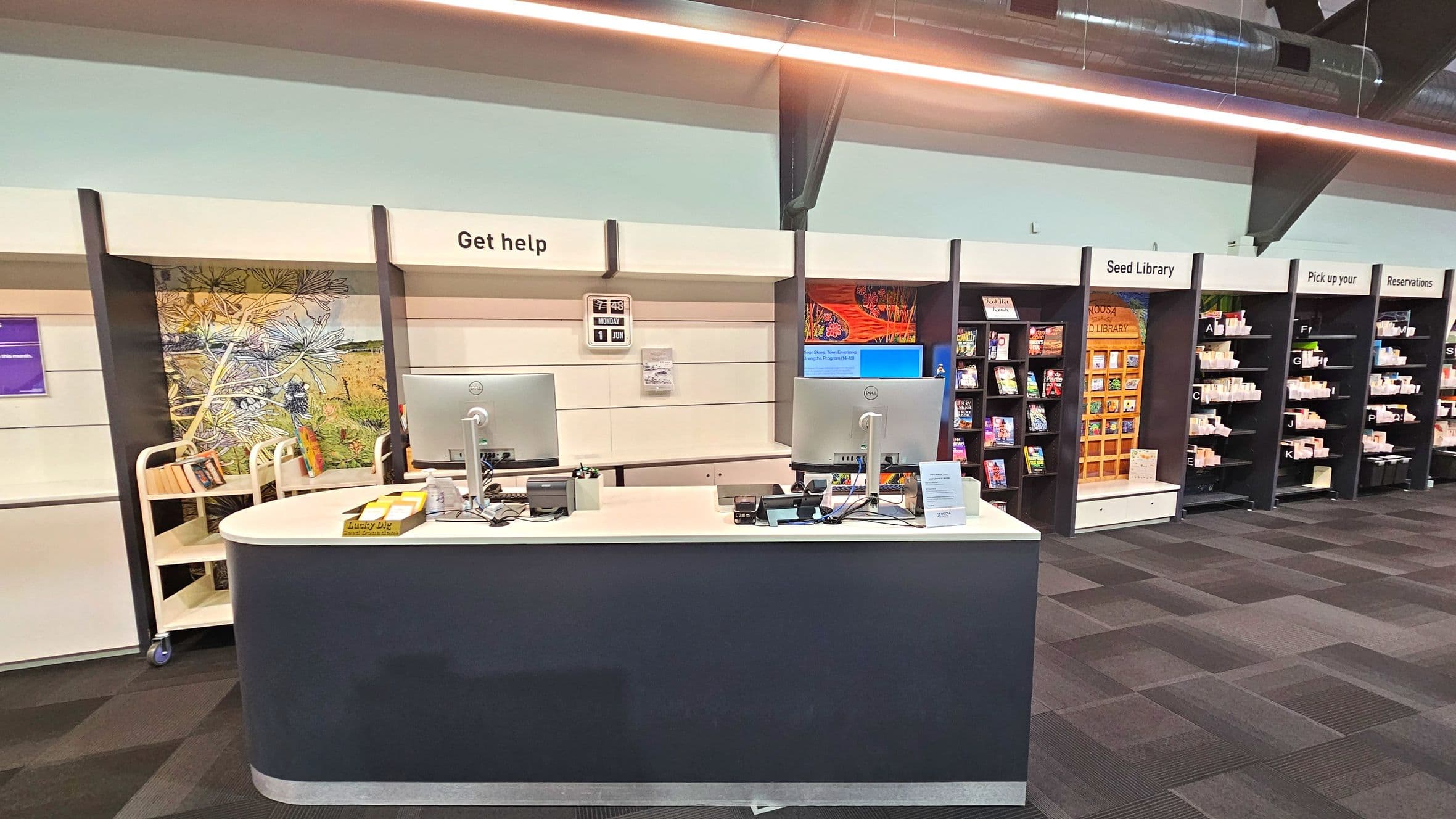 A library help desk with two computer monitors on a curved counter, labelled 'Get help'. Behind the desk, a colourful mural and a trolley with books are visible. To the right, shelves labelled 'Seed Library', 'Pick up your', and 'Reservations' hold various items. The floor is carpeted with a dark checkered pattern, and the ceiling features exposed ductwork and a linear light fixture.