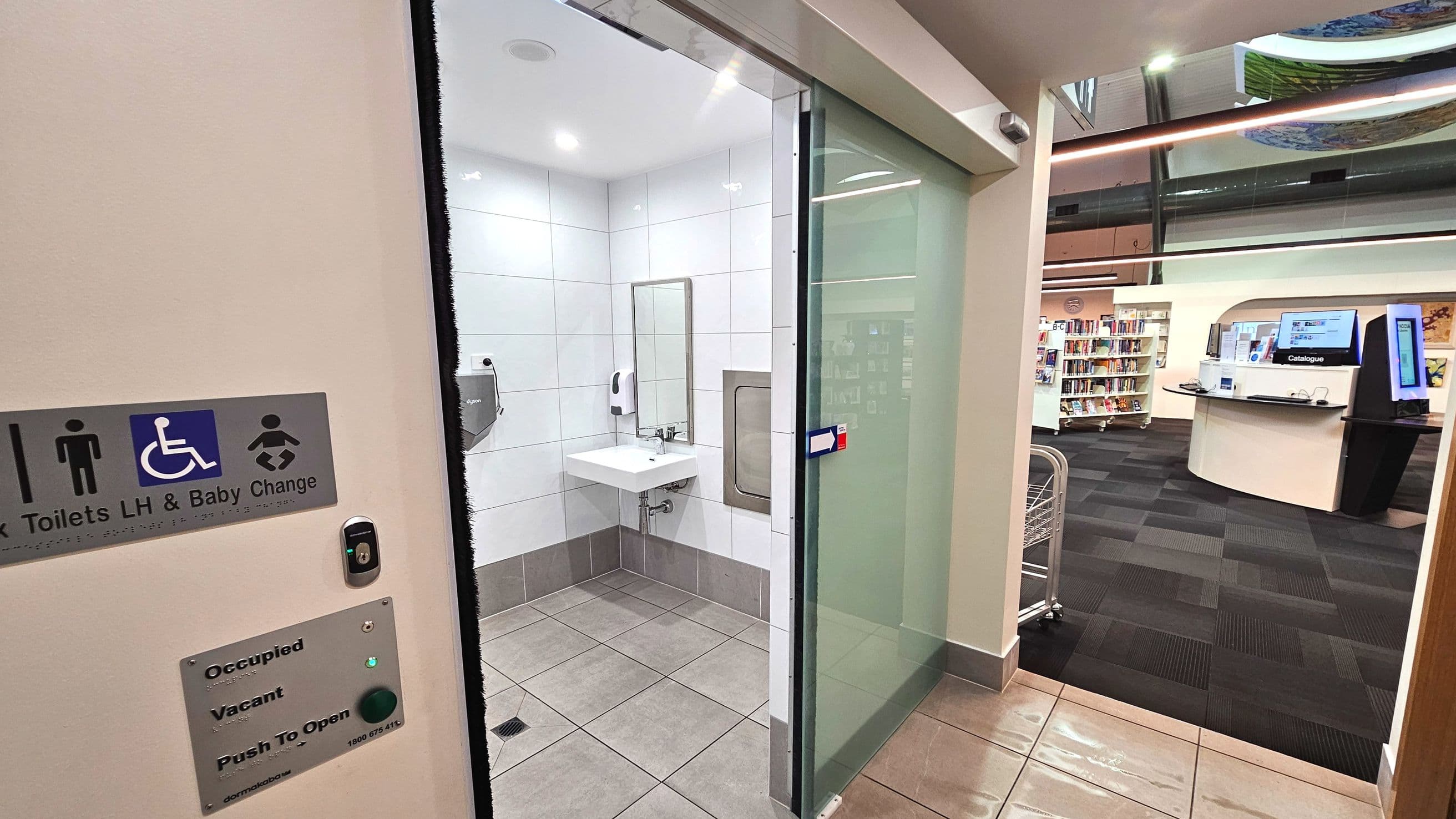 A modern public toilet entrance with a sliding glass door, featuring a sign indicating accessible and baby change facilities. Inside, the room has white tiled walls, a sink with a mirror, and a hand dryer. The floor transitions from grey tiles to dark carpet in the adjacent library area, which includes bookshelves and a digital catalogue station. A push button for door operation is visible on the wall.