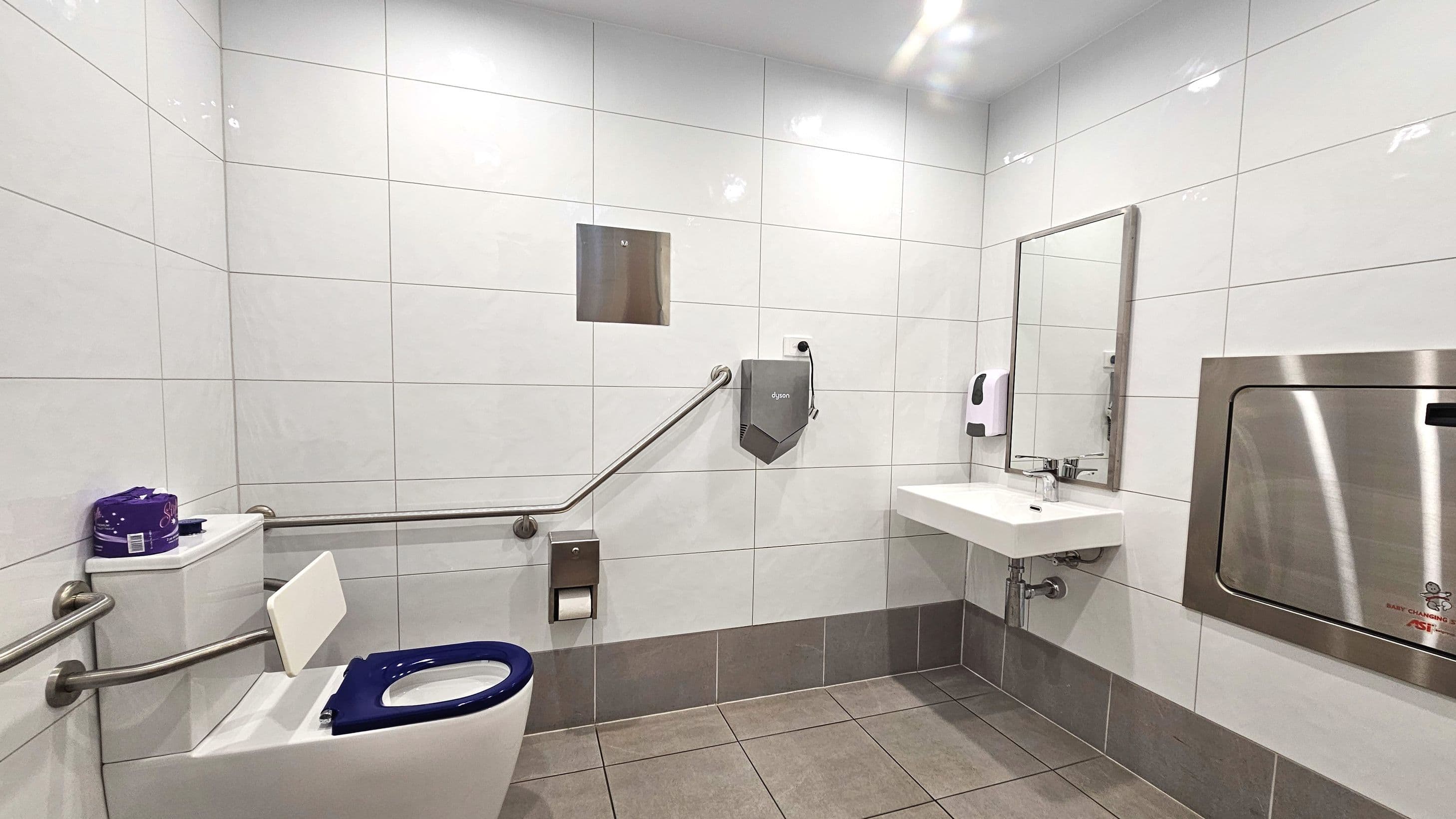 A modern accessible bathroom with white tiled walls and a beige tiled floor. A toilet with a blue seat is equipped with stainless steel grab bars on both sides. A wall-mounted hand dryer and a soap dispenser are near a rectangular sink with a mirror above it. A stainless steel baby changing station is mounted on the wall. A roll of toilet paper is on a holder beside the toilet.