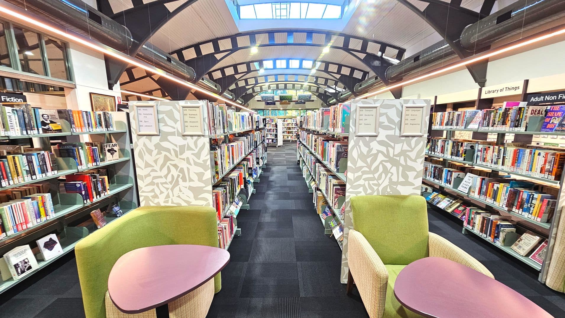 A library interior with high arched ceilings featuring exposed beams and skylights. Two green armchairs with small tables are positioned at the entrance of a central aisle lined with bookshelves. The shelves are filled with books, and signs indicate sections like 'Fiction' and 'Library of Things'. The floor is covered with dark carpet tiles, and the space feels open with ample lighting from overhead fixtures.