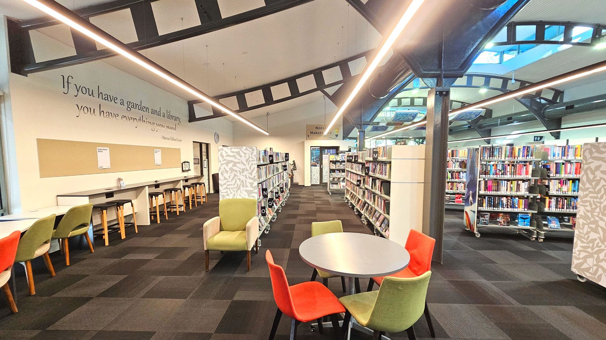 A spacious library interior with a high ceiling supported by black metal beams. The floor is covered with dark patterned carpet tiles. On the left, a long desk with stools is positioned against a wall featuring a quote. In the centre, a round table with colourful chairs is visible. Bookshelves filled with books line the right side, and a sign for the 'Makerspace' is seen in the background.