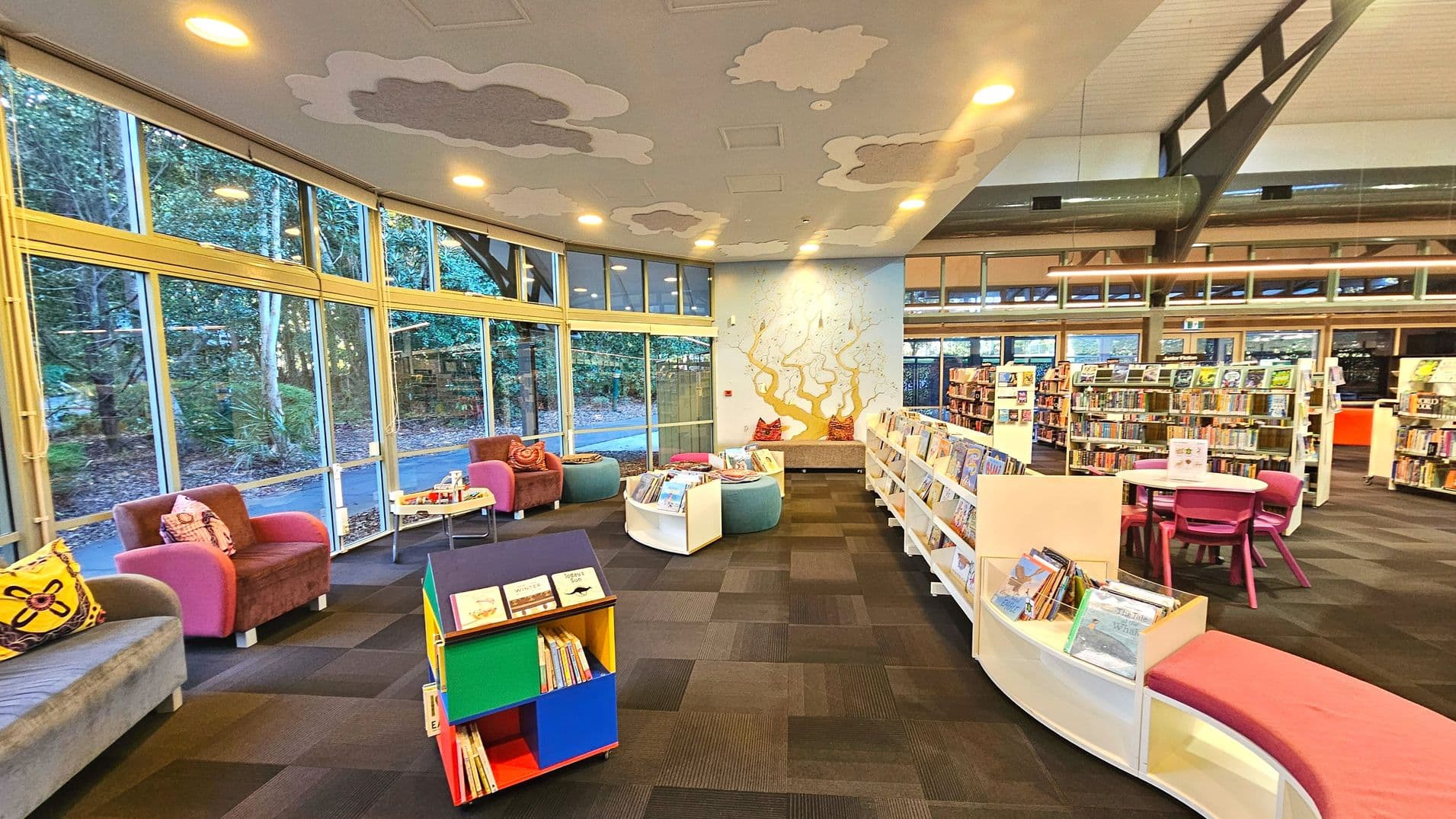 A spacious library interior with large floor-to-ceiling windows on the left, offering a view of lush greenery outside. The room features colourful seating, including pink and purple armchairs and a curved bench with cushions. Bookshelves are arranged in a semi-circle, filled with books. A round table with pink chairs is visible on the right. The ceiling has cloud-shaped decorations, and a wall mural of a tree adds artistic flair.