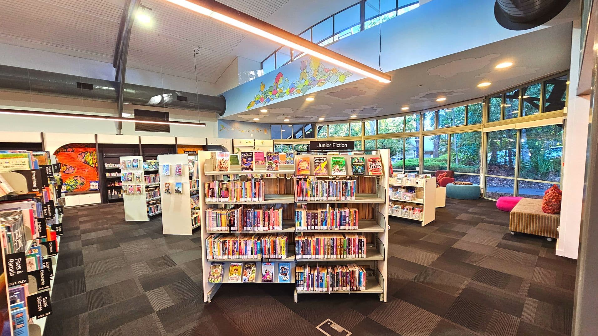 A library interior featuring a central bookshelf labelled 'Junior Fiction' filled with colourful children's books. The space has a high ceiling with exposed ductwork and a large mural above. To the right, large windows offer views of greenery outside. The floor is covered with dark, patterned carpet tiles. There are cushioned seating areas with vibrant colours near the windows, creating a welcoming reading nook.