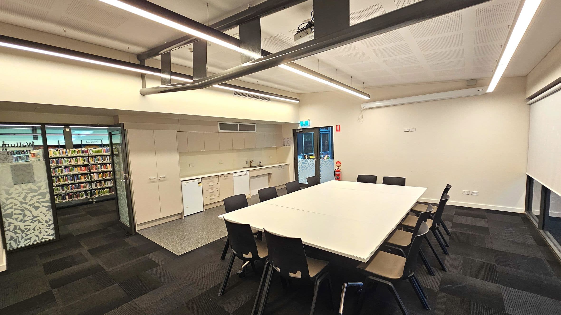 A modern meeting room with a large rectangular table surrounded by black chairs on a dark carpeted floor. The room features a kitchenette with white cabinets, a sink, and a small fridge along one wall. A glass door with patterned frosting leads to a library area filled with bookshelves. Overhead, linear fluorescent lights and exposed ductwork are visible. A fire extinguisher is positioned near the exit door.