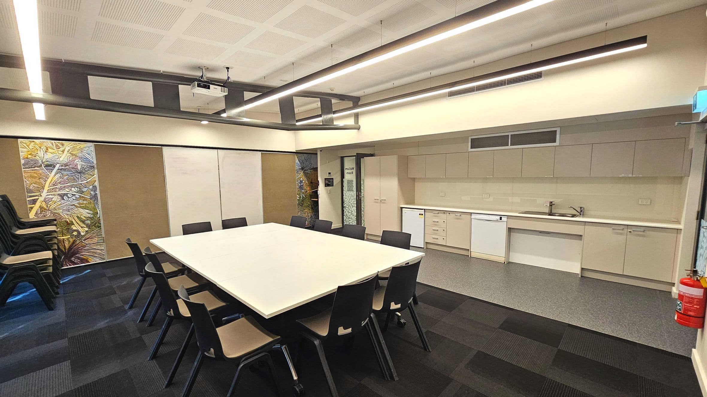 A modern meeting room with a large rectangular table surrounded by black chairs. The room features a kitchenette along one wall with beige cabinets, a sink, and a dishwasher. A stack of additional chairs is in the corner. The ceiling has recessed lighting and a projector. The floor transitions from dark carpet to light vinyl near the kitchenette. A fire extinguisher is mounted on the wall.