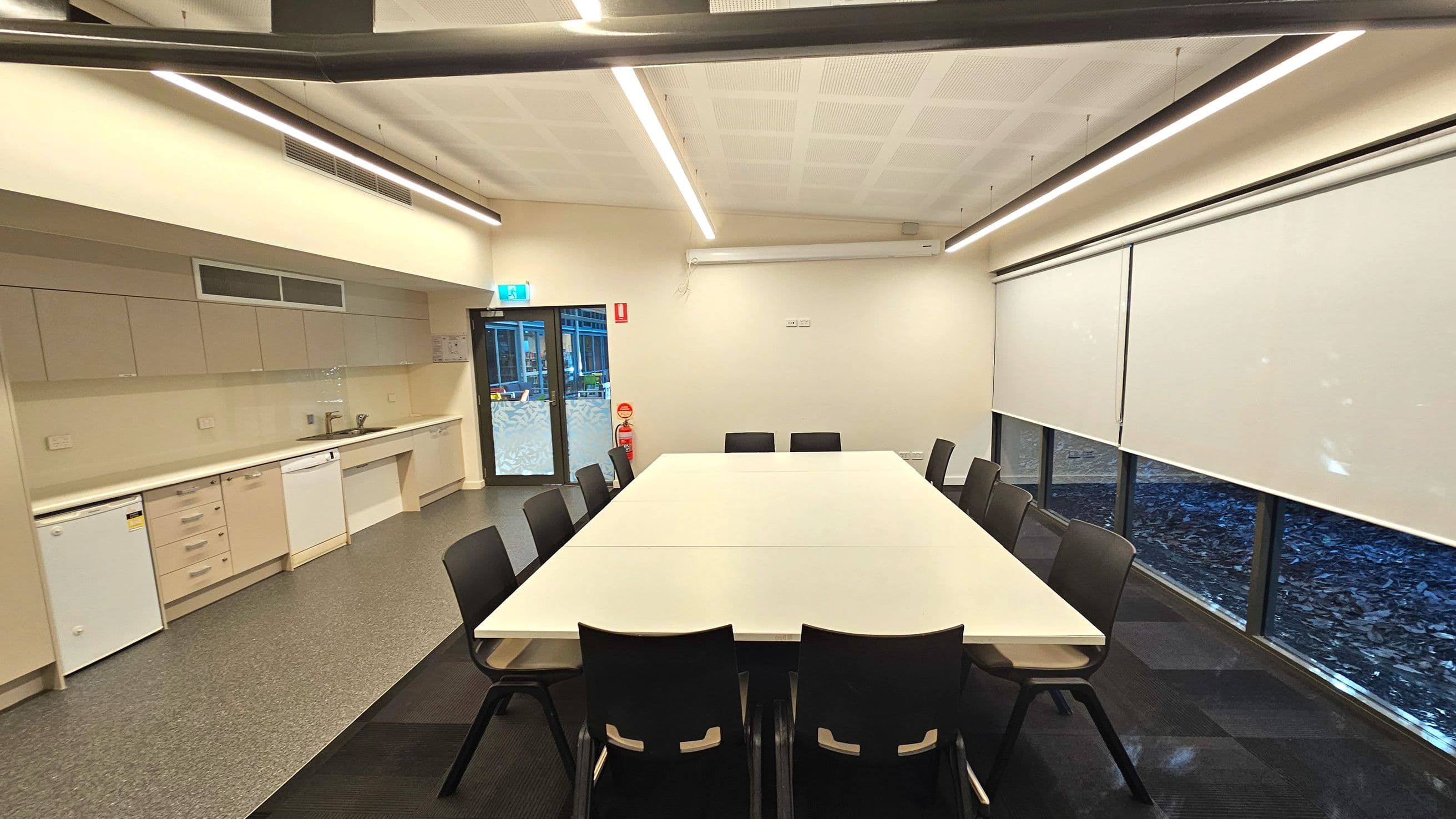 A modern meeting room with a large rectangular white table surrounded by black chairs. On the left, a kitchenette features white cabinets, a sink, and appliances including a dishwasher and mini-fridge. The floor transitions from grey tiles in the kitchenette to dark carpet near the table. Large windows with white roller blinds line the right wall. A glass door with an exit sign is at the back, next to a fire extinguisher.